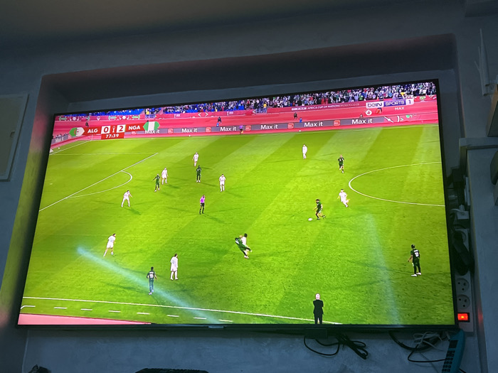 A television screen shows a soccer game between Algeria (ALG) and Nigeria (NGA). The score is 0-2 in favor of Nigeria at 77:39 minutes into the match. The game is being broadcast on beIN SPORTS 1. The pitch is green with white markings, and players in white and green jerseys are visible. The field is surrounded by spectators. Multiple "Max it" banners are also visible. A man in a black suit stands near the bottom of the screen.