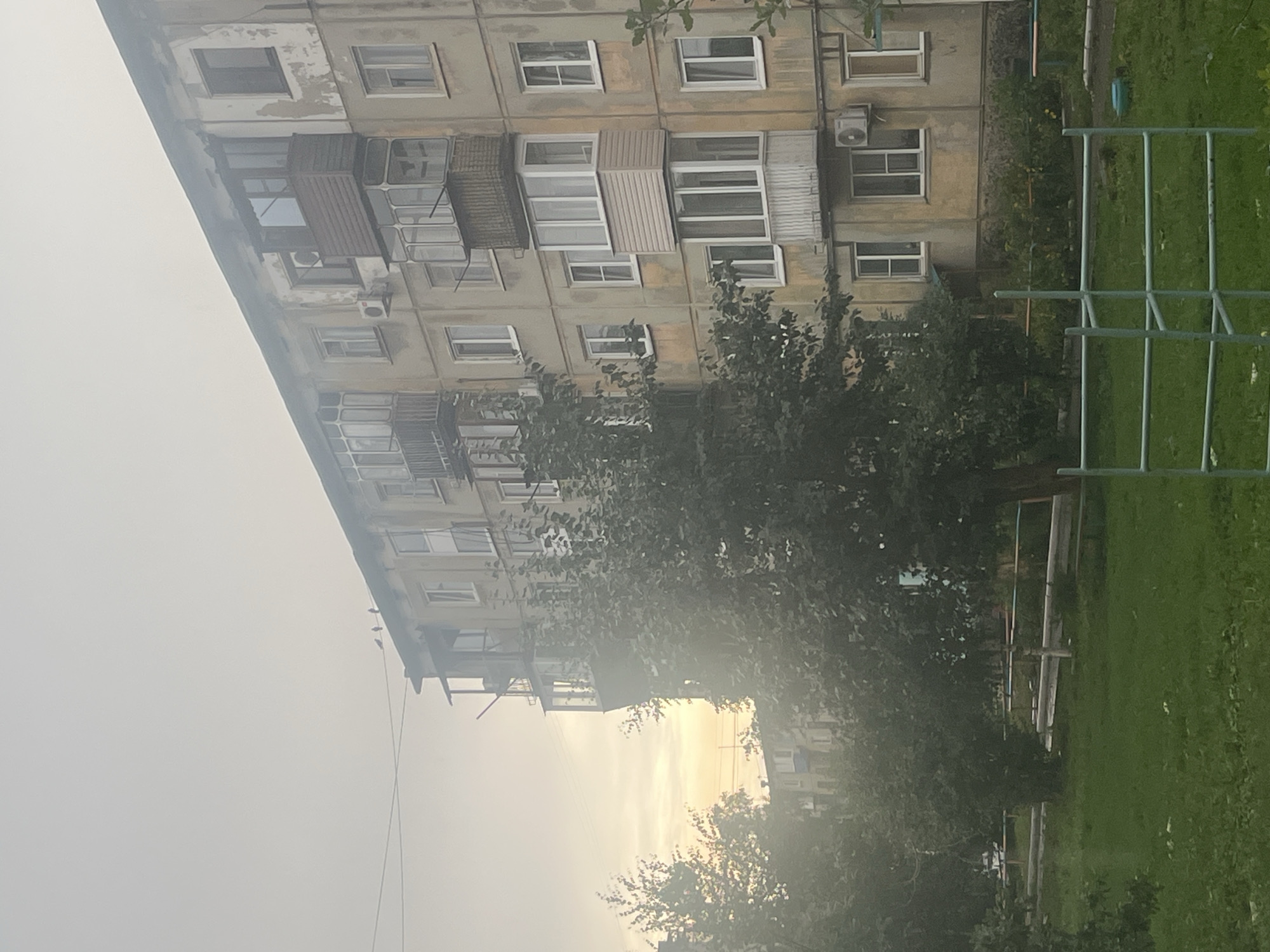 The image captures a residential building, likely an apartment complex, in the city of Oskemen, Kazakhstan. The building is weathered, with peeling paint and visible signs of age. It features multiple stories, with numerous windows and balconies. Some balconies are enclosed with glass, while others are open. An air conditioning unit is visible on the facade. In front of the building is a grassy area with trees, a paved path, and some playground equipment, suggesting a residential courtyard. The sky is overcast, with a soft light indicating either early morning or late afternoon. A few birds are perched on a wire in the upper portion of the image. The overall impression is of a typical urban residential setting in the region.
