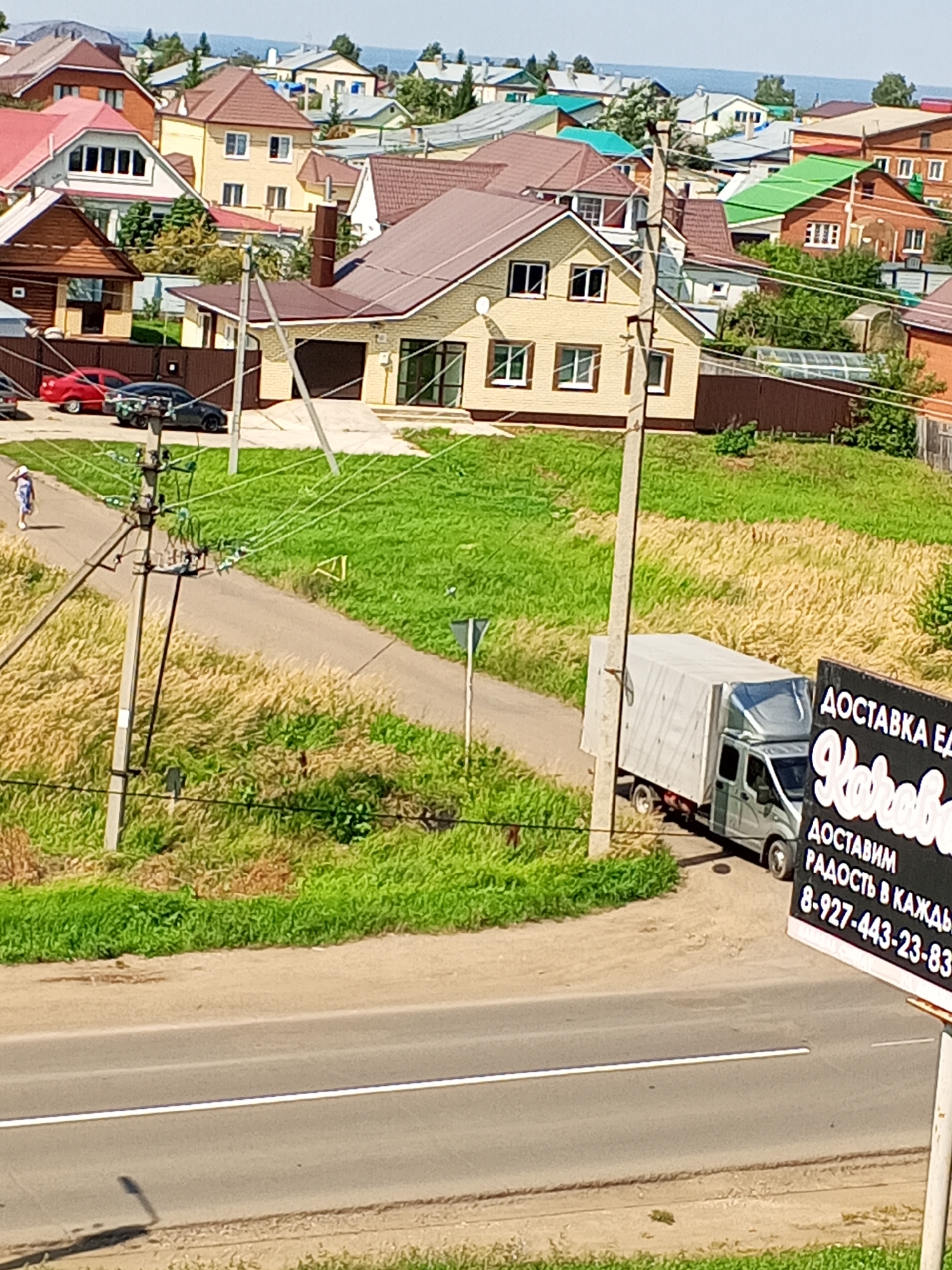 The image depicts a residential area with multiple houses of varying architectural styles, predominantly featuring pitched roofs covered in brown and red tiles, and some with green roofs. The houses are situated on a gentle slope, with some appearing to be under construction or recently built, indicated by exposed construction materials like brickwork. A paved road runs through the scene, bordered by green grass and tall, dry grass or reeds.
In the mid-ground, a light-colored delivery truck with a white canvas cover is parked on the side of the road, facing away from the camera. Beside the truck, a dark sign with white lettering is visible, displaying Russian text and a phone number. The sign appears to be advertising a delivery service.
A person wearing a white hat and blue and white clothing is walking on the road in the upper left portion of the image. Electrical poles and wires are prominent elements in the scene, crisscrossing the sky and landscape, suggesting an urban or suburban setting. The overall environment is sunny, with clear blue skies visible in the background.
While the image is visually rich, specific landmarks that definitively place it in Chistopol, Russia are not immediately apparent from the visual cues alone. However, the presence of Russian text on the sign strongly suggests a Russian location. The architecture of the houses, characterized by their multi-story design and pitched roofs, is consistent with residential construction found in many Russian towns and cities. The overall landscape, with its rolling terrain and vegetation, is also typical of rural or semi-rural areas within Russia.