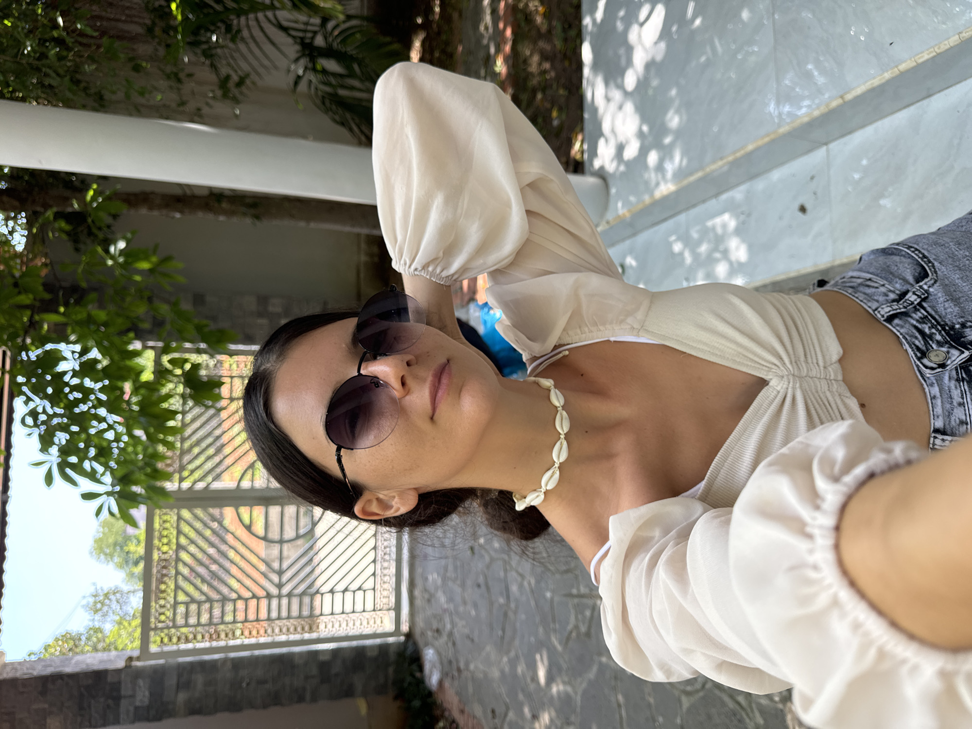 A young woman is taking a selfie outdoors in Mui Ne, Vietnam. She is wearing a cream-colored ruched top with voluminous sheer sleeves, a white bikini top underneath, and grey denim shorts. She has on round, dark sunglasses and a choker necklace made of small white shells. Her right arm is raised, holding the phone, and her left hand is resting on the back of her neck.
The background shows an outdoor setting with a paved walkway and a white pillar on the right side. Beyond the walkway is a decorative metal gate with a brick wall and greenery visible through it. Trees and lush foliage are present in the background, with dappled sunlight filtering through the leaves. The overall impression is a sunny, casual, outdoor environment, likely in a residential or resort area of Mui Ne.