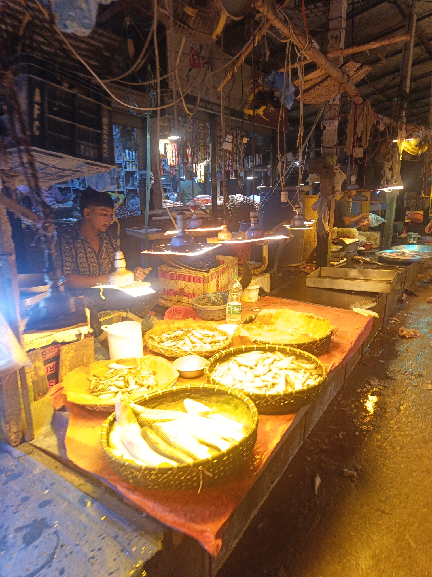 This is a photograph taken at a fish market, likely in Dhaka, Bangladesh, given the visual cues. The scene is bustling with activity, primarily centered around the sale of fresh fish.
In the foreground, a vibrant display of fish is laid out on an orange cloth-covered table. Several woven baskets are filled with various types of fish. One basket prominently showcases larger, elongated fish, while another holds smaller, more numerous fish. A small metal bowl is placed next to one of the baskets. Adjacent to these, a large round tray also contains a pile of smaller fish. A white plastic bucket sits to the left of the fish display.
To the left, a young man, wearing a patterned short-sleeved shirt and dark trousers, is seated and appears to be preparing or arranging fish. He is illuminated by a bright light, possibly a lamp, positioned above his workspace. Behind him, shelves are visible, stocked with various items, and some signage is present, though illegible.
The market environment is dimly lit, with overhead lights creating a warm, yellow glow. The ceiling is constructed from bamboo or wood, with a complex network of wires, pipes, and hanging materials, suggesting a rustic and functional setup. The floor appears wet and somewhat grimy, reflecting the ambient light, indicating a typical market condition. Further back, other vendors and customers can be vaguely seen, engaged in their transactions. The overall impression is that of a lively, traditional marketplace, likely operating during evening hours given the artificial lighting.