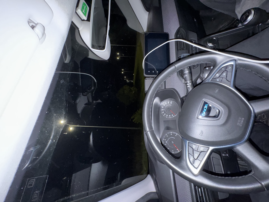 The image presents a nighttime view from within a Dacia passenger vehicle, specifically from the driver's perspective. The black steering wheel with the Dacia logo and integrated controls dominates the foreground. Behind it, the instrument cluster displays analog speedometer and tachometer dials, both indicating zero. A central touchscreen infotainment system is mounted on the dashboard. A white cable extends from the upper interior, across the infotainment screen, and descends toward the passenger footwell. A manual gear shift lever is visible to the right of the center console. Affixed to the upper windshield near the rearview mirror is a green Via Verde electronic toll transponder. Through the windshield, a dark outdoor environment is visible, illuminated by a series of amber streetlights lining a road or open area that recedes into the distance. Sparse vegetation is discernible beyond the immediate illuminated zone. The scene is located in Mirandela, Portugal.
