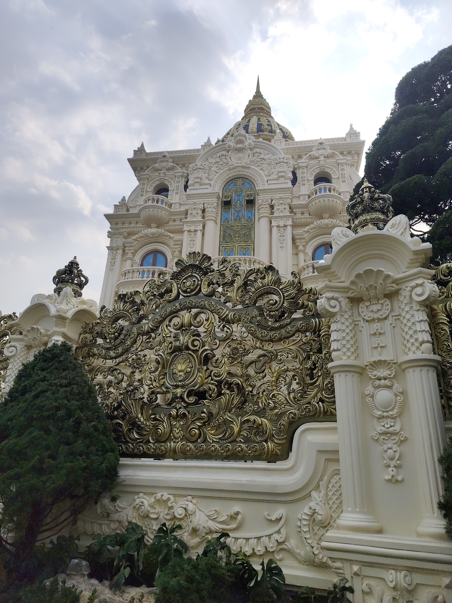 The image shows a low-angle view of a building with ornate, gilded details, presumably a palace or temple. The structure is predominantly white, but the gate is elaborately crafted with gold-colored filigree. The building itself features a dome and classical architectural elements like pillars and arches. There are trees and plants surrounding the base of the structure, suggesting a garden setting. The sky is overcast. The location is in Da Lat, Vietnam, and the building is associated with the Linh Phong Buddhist Temple.