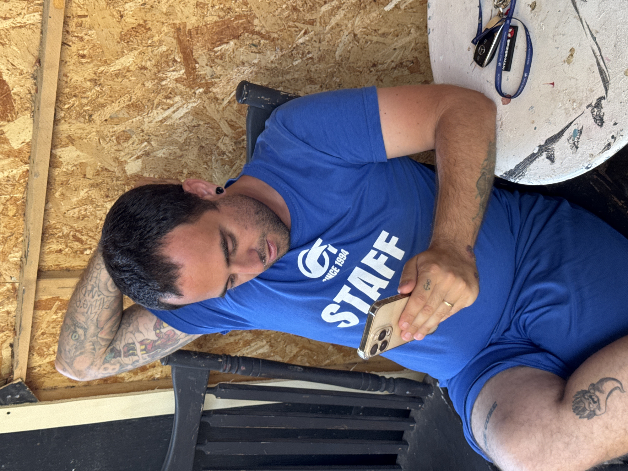 A man with dark hair, a beard, and tattoos on his arms is sitting on a chair. He is wearing a blue t-shirt and blue shorts. He is holding a smartphone in his hands and looking at it. On the table next to him are a set of keys. The background is a wooden wall and a black chair. The scene appears to be outdoors, possibly on a porch or patio. The image is taken in Isola delle Femmine, Italy.