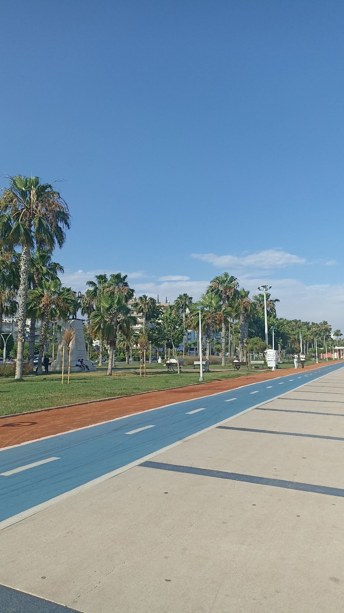 This media file captures a serene outdoor scene in Yenişehir, Turkey, under a clear blue sky with a few wispy clouds. The time of day appears to be late morning or early afternoon, with bright sunlight casting distinct shadows, suggesting pleasant weather.
The foreground is dominated by a paved walkway with a distinct blue lane marked with white dashes, likely a designated path for cycling or rollerblading. Adjacent to this blue lane is an orange-colored track, possibly for running, and a strip of green grass. Palm trees are a prominent feature throughout the scene, lining the park area.
In the middle ground, a park area features more palm trees, grassy lawns, and several individuals enjoying the outdoors. Some people are seated on benches, while others are walking or standing. A prominent monument or statue stands on a pedestal, with a few people gathered near it. Buildings can be seen in the background, suggesting an urban or semi-urban setting. Streetlights are also visible, indicating the area is well-equipped for public use.
Further along the walkway, more people are seen strolling, and the path continues into the distance, lined with palm trees and more parkland. The overall mood of the image is peaceful and inviting, conveying a sense of leisure and community. No visible text is present in the image.