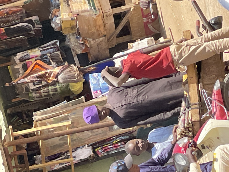 An outdoor market scene in Samaru, Nigeria, features five individuals gathered around a wooden stall under bright daylight. A central adult male, wearing a purple skullcap and a dark brown long-sleeved tunic, stands looking left with a slight smile. To his immediate left, a child in a white and light blue shirt also looks left. To the central male's right, a child in a red polo shirt and tan pants is seen from behind, with his right hand on the back of his head. In the lower left foreground, a person partially obscured by a dark cap is positioned on a red and white scooter/motorcycle, with hands on the handlebars. A wooden display platform in front of the central male and the child in white holds bundles of cut sugarcane. The background stall exhibits various packaged hair extensions, fabrics, stacked cardboard boxes, a hanging patterned scarf, and a light-colored wig form. The ground is dry earth.