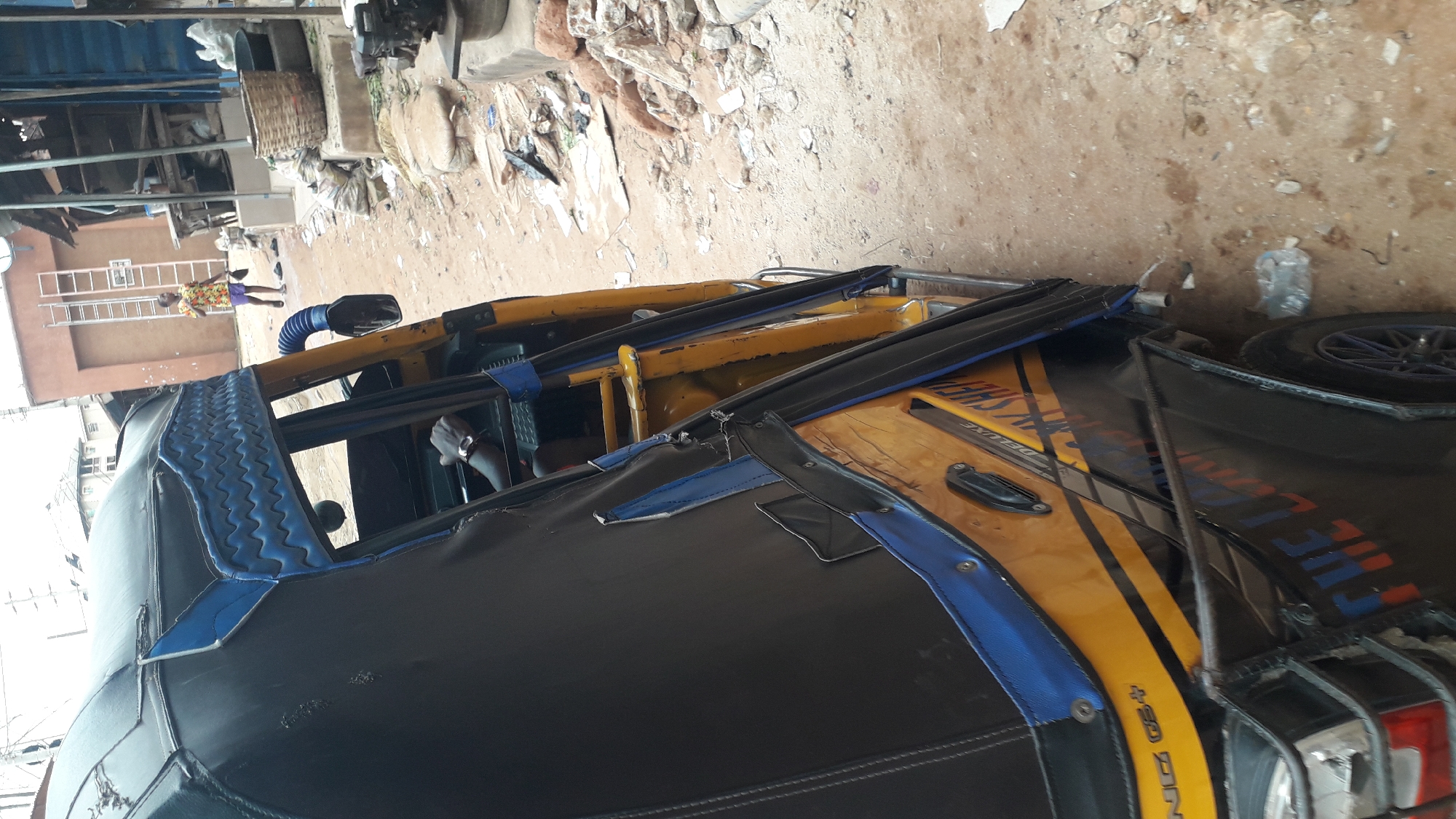 An overhead shot shows the side of a yellow and black motorized rickshaw parked on a dirt road. The rickshaw has a blue and black striped trim along the top. A person is partially visible in the driver's seat, wearing a watch. In the background, a young person in a colorful shirt and shorts walks away from a building with a ladder. The environment appears to be a market or a residential area with debris and scattered items on the ground.