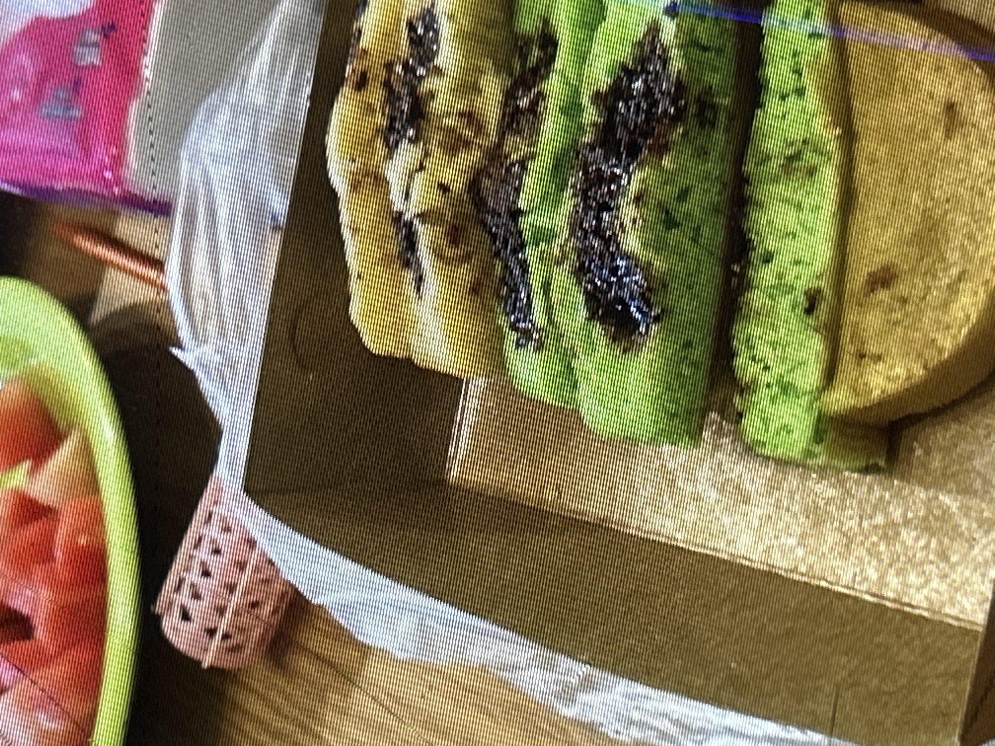 The image shows a close-up of several layers of brightly colored cake, likely Kue Lapis, a traditional Indonesian layered cake. The cake is arranged in a cardboard box, with distinct green and yellow layers. Some dark specks, possibly chocolate or a similar flavoring, are visible on the cake's surface. In the background, out of focus, there is a plate with sliced red fruit, possibly watermelon, and a pink package. The overall setting appears to be indoors, on a textured surface that could be a table. There are no people visible in the image, and the focus is entirely on the food items. Without further context, it is not possible to determine the specific location within Depok, Indonesia.