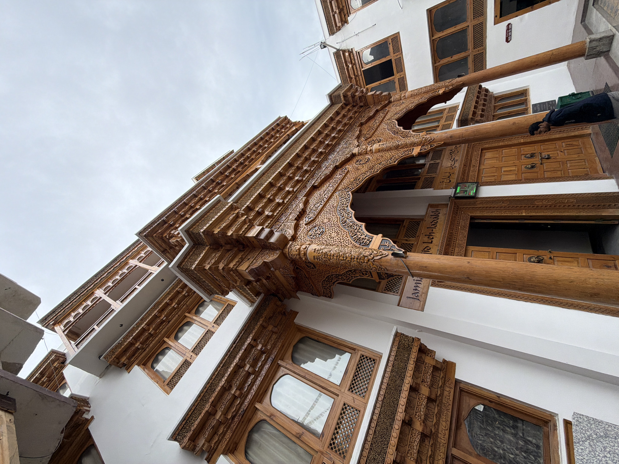 A low-angle, wide shot captures the intricate wooden facade of the Jama Masjid in Leh, India. The building features white walls adorned with elaborate, dark wood carvings around windows and the entrance. A large, ornate wooden archway frames double wooden doors, each with round brass knockers. A man in a dark jacket and gray pants stands to the right of the entrance, facing away from the camera and towards the building. Above the entrance, a wooden sign reads "Jama Masjid Leh-Ladakh". The sky above is overcast and pale.