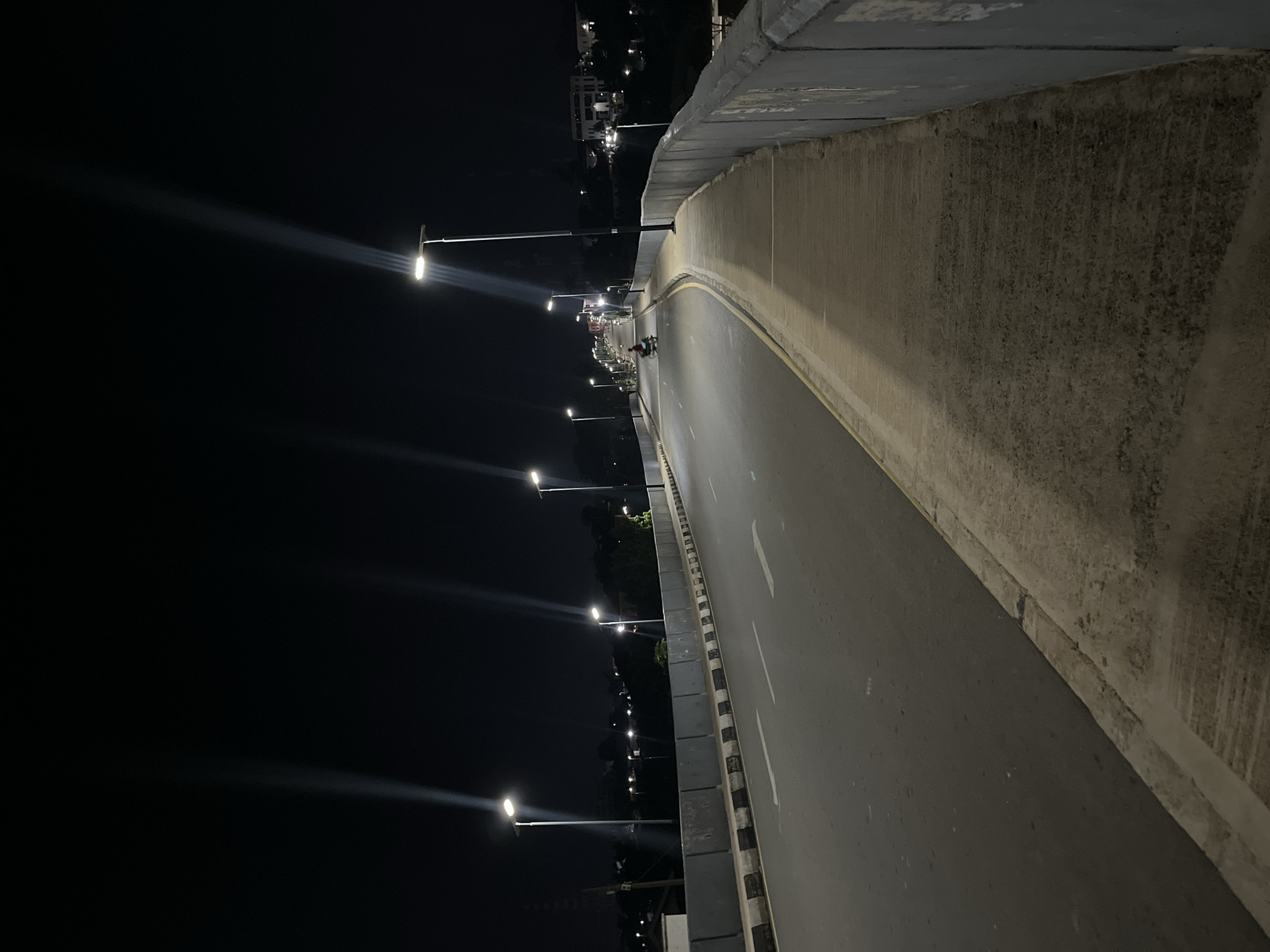 The image shows a bridge at night in Jos, Nigeria. A row of streetlights illuminates the bridge roadway. A cyclist and a pedestrian are visible in the distance, moving along the bridge. The bridge has concrete barriers on either side. The sky is dark. The bridge surface appears to be asphalt.