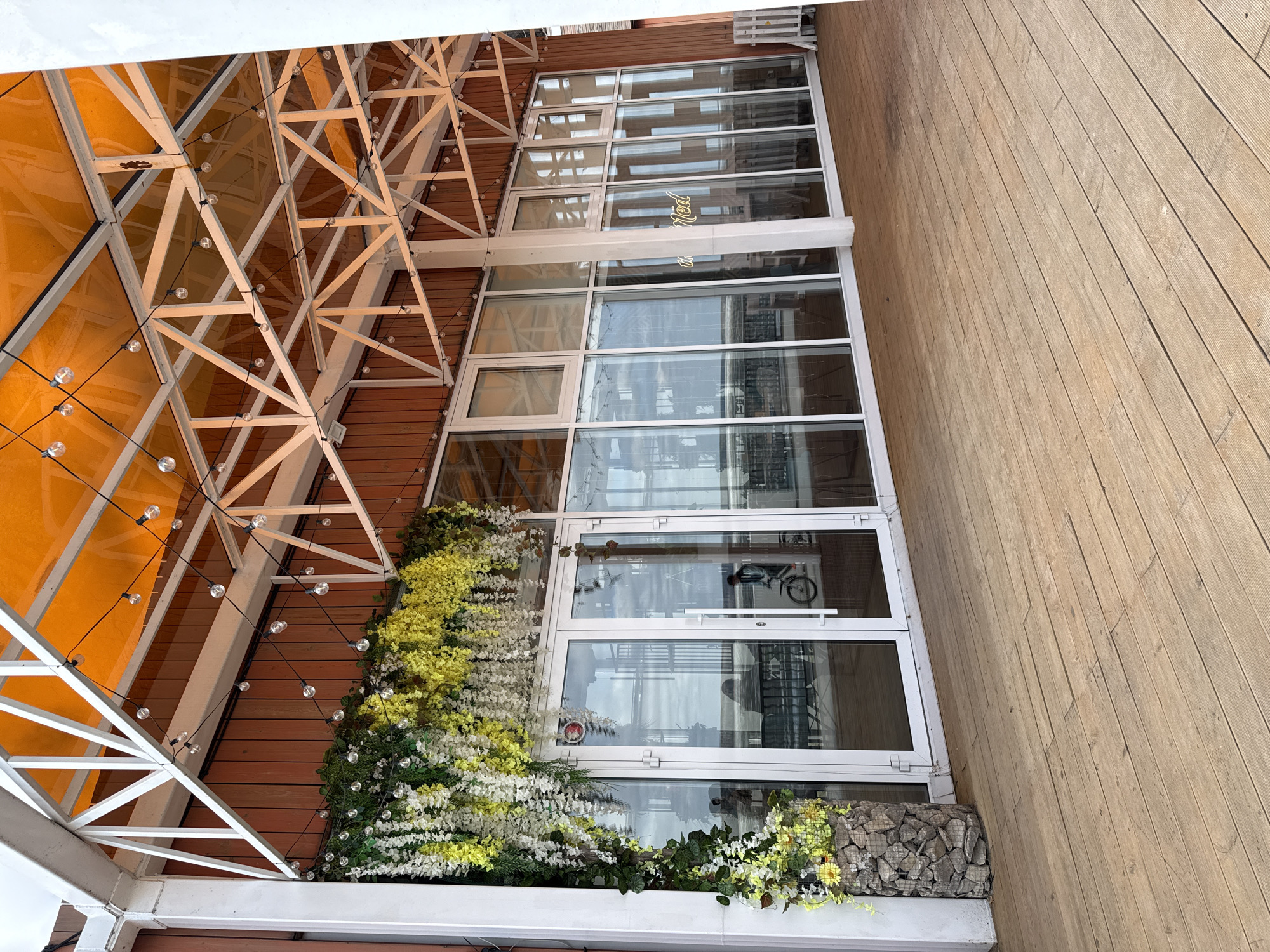 The image depicts a building's facade in Chaikovsky, Russia. The building has a glass and white-framed window wall adorned with floral arrangements. A metal frame canopy with a partially translucent orange roof covers the top section. String lights are hung along the metal frame. The ground adjacent to the building is a wooden deck. A bicycle is visible through the glass window. There is no one present.