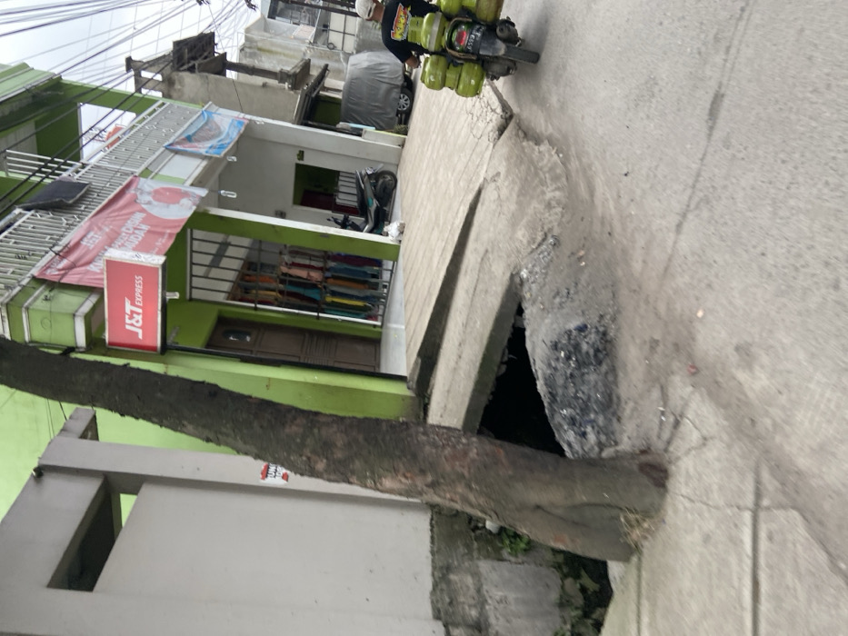 The image shows a street in Margaasih, Indonesia, at the coordinates -6.954 latitude and 107.551 longitude. A large tree trunk is located in the foreground and is growing at the edge of the street. The tree trunk is partially covering a large hole in the street, which is filled with dirt and debris. The road is paved with concrete and there is a small amount of debris and dirt scattered on the surface. There is a small building with a green facade and a sign that reads "J&T Express" to the left of the image. A person is riding a motorcycle with multiple green cylinders attached to the back. The person is wearing a black shirt and a white helmet. There is a small amount of pedestrian traffic in the background. The sky is overcast and there are power lines running across the street. The building has a small wooden door and a metal gate with colorful fabrics hanging inside the building. There is a small white cat visible on the sidewalk. A motorcycle with a rider is traveling away from the camera towards the left of the image. The motorcycle is carrying multiple green cylinders. The environment is urban and there are several buildings in the background. The sidewalk is cracked and uneven and there is a small drain in the foreground. The street is fairly narrow and the overall scene is one of a typical urban street in a developing country.