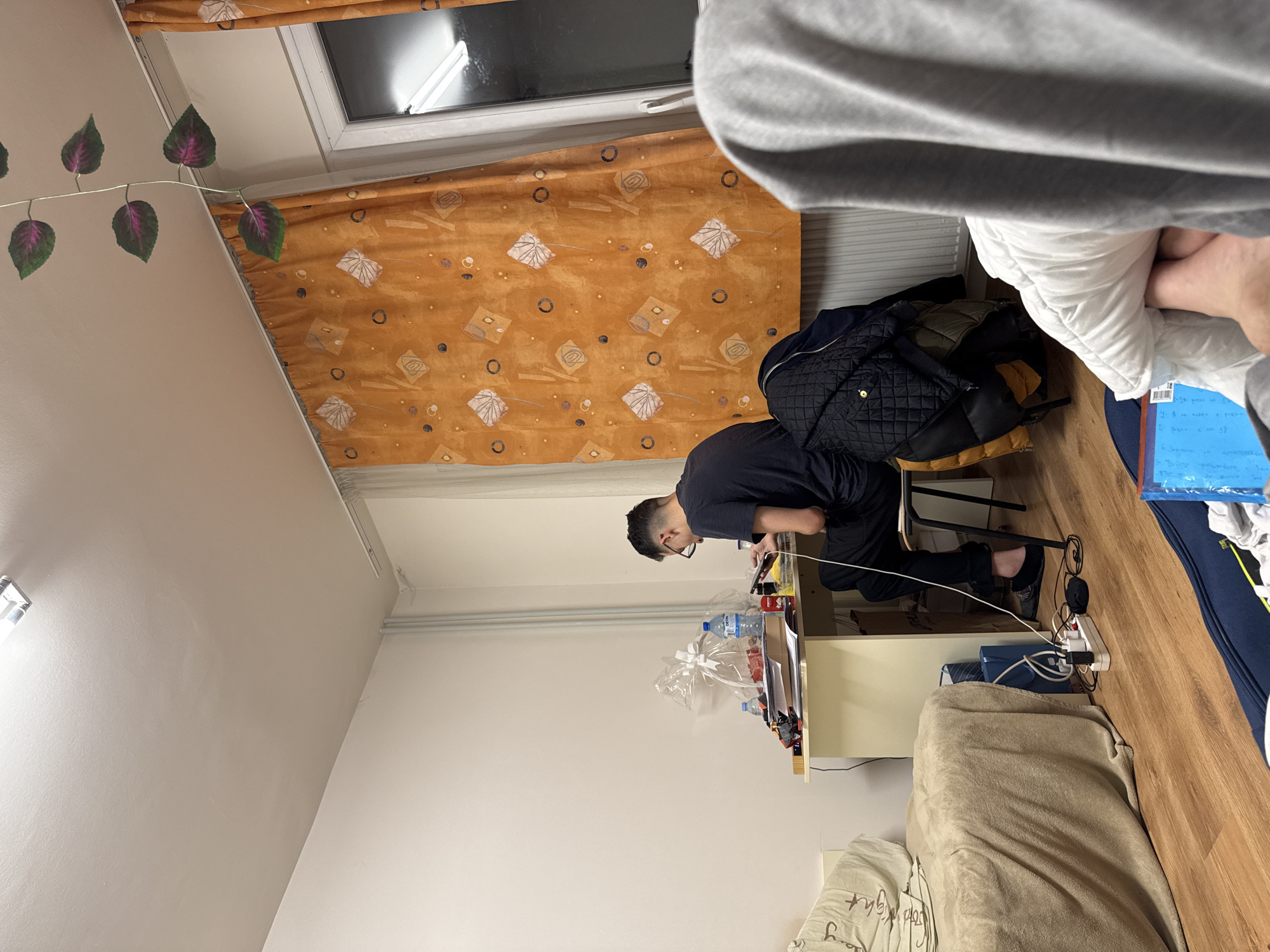 A rotated indoor shot depicts a person in a black shirt and pants sitting at a desk with multiple items. Water bottles, boxes, and what appear to be electronic devices are on the desk. The person is looking down at the items on the desk, possibly interacting with them. A window with an orange curtain is visible behind the person. A bed with a tan blanket is partially visible in the foreground. Power cords and a power strip are on the floor. The presence of Bulgarian text on the blue paper suggests the location is likely Sofia, Bulgaria. The shot is obscured by someone else in the foreground.