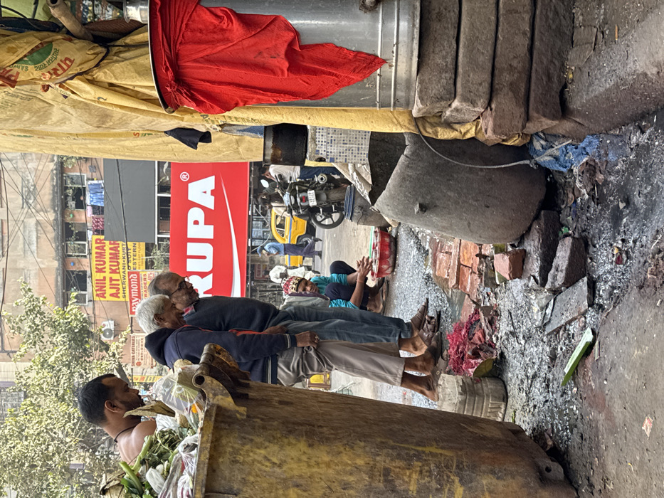 An urban street scene shows two elderly men seated on the ground. Another man stands beside a dumpster filled with green waste. A woman sits on the ground clapping. Overhead, a red "RUPA" sign is visible, along with store signs below. A yellow auto-rickshaw and motorbike are parked nearby. The ground is littered with debris, including bricks and broken concrete. Above, tarps and a red cloth are draped over pipes and concrete blocks.