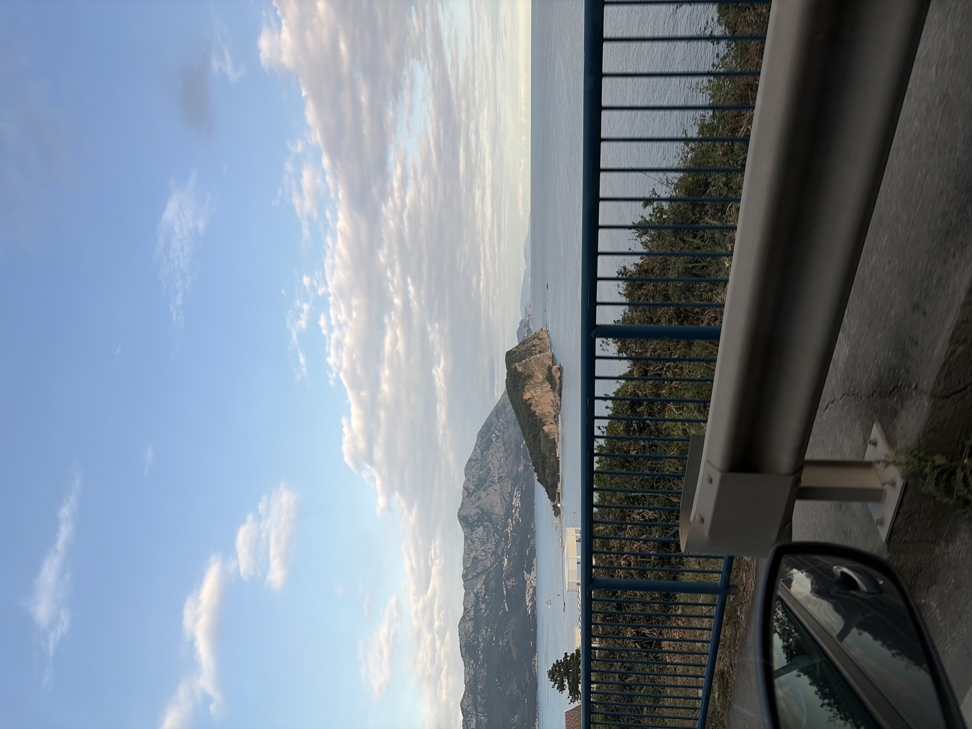 The image shows a view from a vehicle traveling along a coastal road, likely near Budva, Montenegro. In the foreground, a gray guardrail and the side mirror of the vehicle are visible. The background features a panoramic view of the Adriatic Sea with a mountain range extending into the water. The sky is partly cloudy with a gradient of blue and white. The scene appears to be captured during daylight, possibly late afternoon, based on the light.