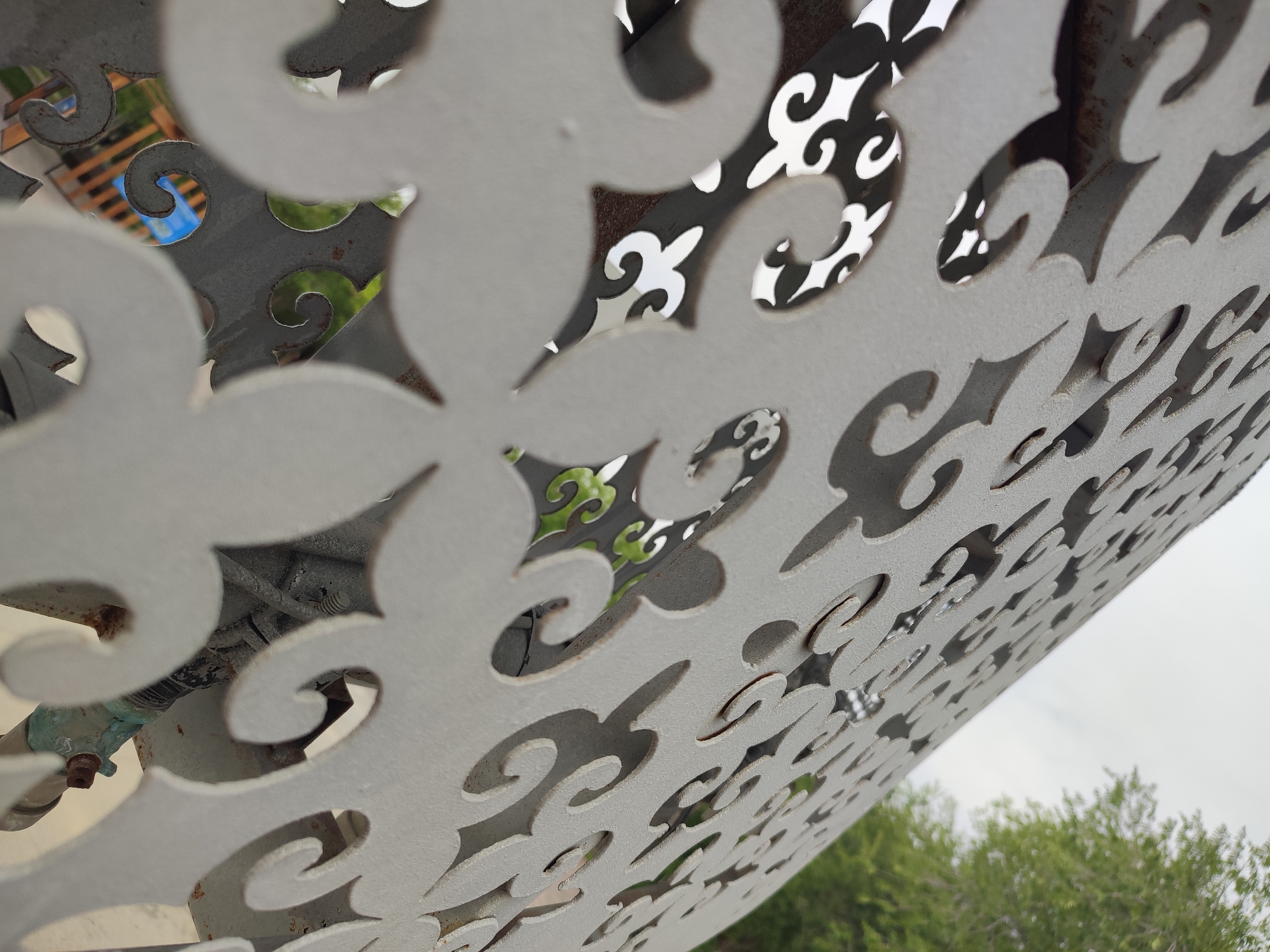 A close-up, low-angle shot reveals a weathered, white metal screen with intricate, swirling cutouts. The screen is positioned outdoors, with a blurred background showing greenery and a cloudy sky, indicative of Qonaev, Kazakhstan. The screen itself shows signs of age, with rust spots visible along the edges of the cutouts and on some supporting metal structures. Through the openings of the screen, hints of darker metalwork, possibly part of a bench or table, and some blue and green elements are visible. The foreground is dominated by the patterned metalwork, emphasizing its texture and decorative design.