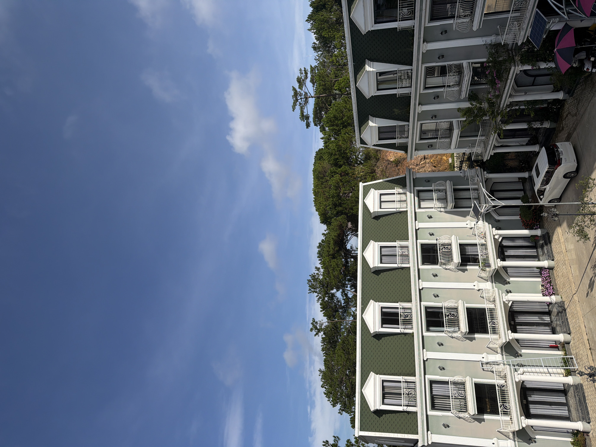 Two adjoining light gray buildings with mansard green roofs, featuring numerous dormer windows and white-railed balconies, are situated in Xuân Trường - Đà Lạt, Vietnam. The buildings exhibit neoclassical architectural elements, including Corinthian columns and decorative molding. In front of the buildings, a white minivan is parked on a paved area, alongside a lamppost with solar panels. To the right, a pink and black umbrella is visible. Lush green pine trees form a backdrop, with a rocky hillside peeking through on the right side of the frame. The sky is a clear blue with scattered white clouds.