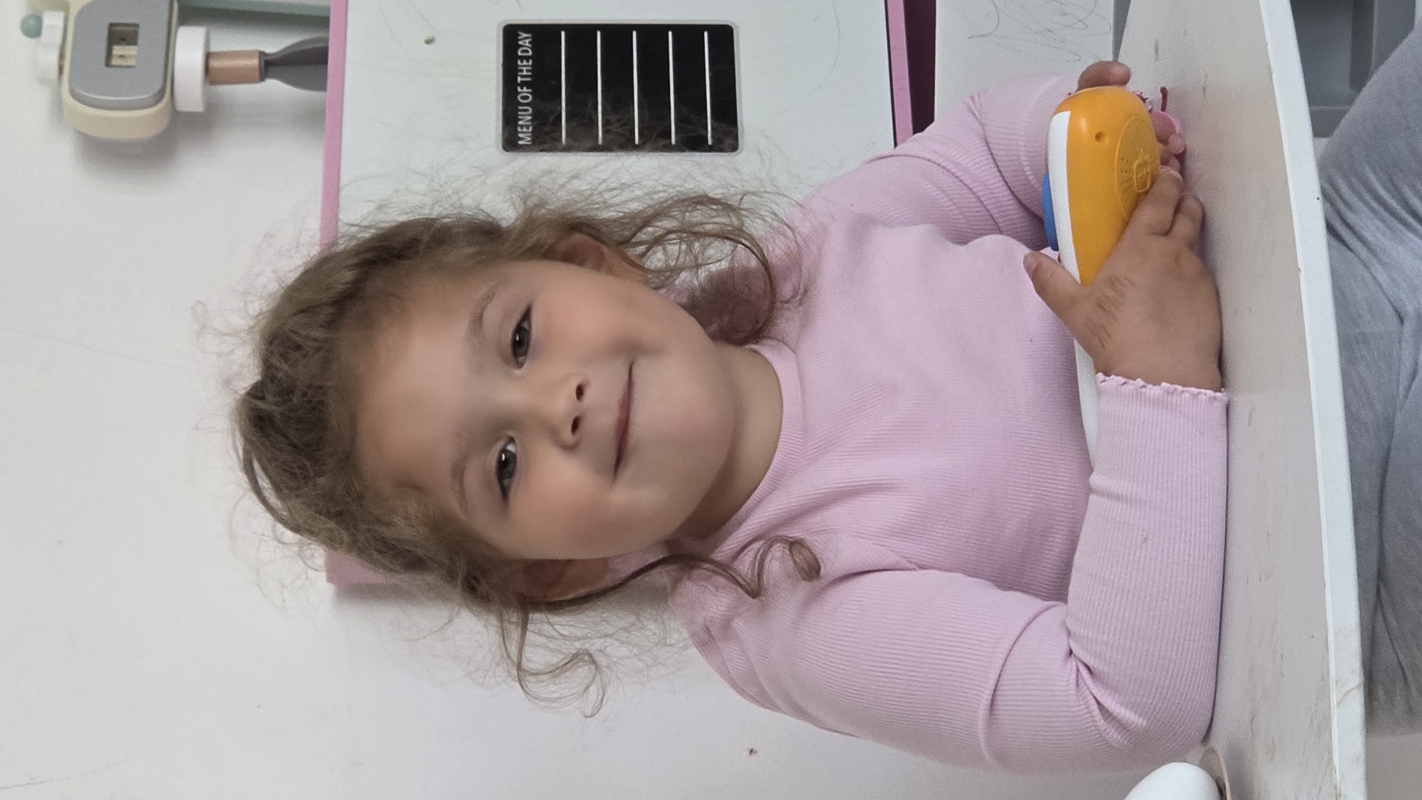A young girl with curly brown hair and brown eyes, wearing a long-sleeved pink shirt, is smiling and looking at the camera. She is holding a yellow and blue toy in her hands. To her left, there is a white board with a "MENU OF THE DAY" sign. The location appears to be indoors. There are no indicators to place this photo in the city of London, United Kingdom.