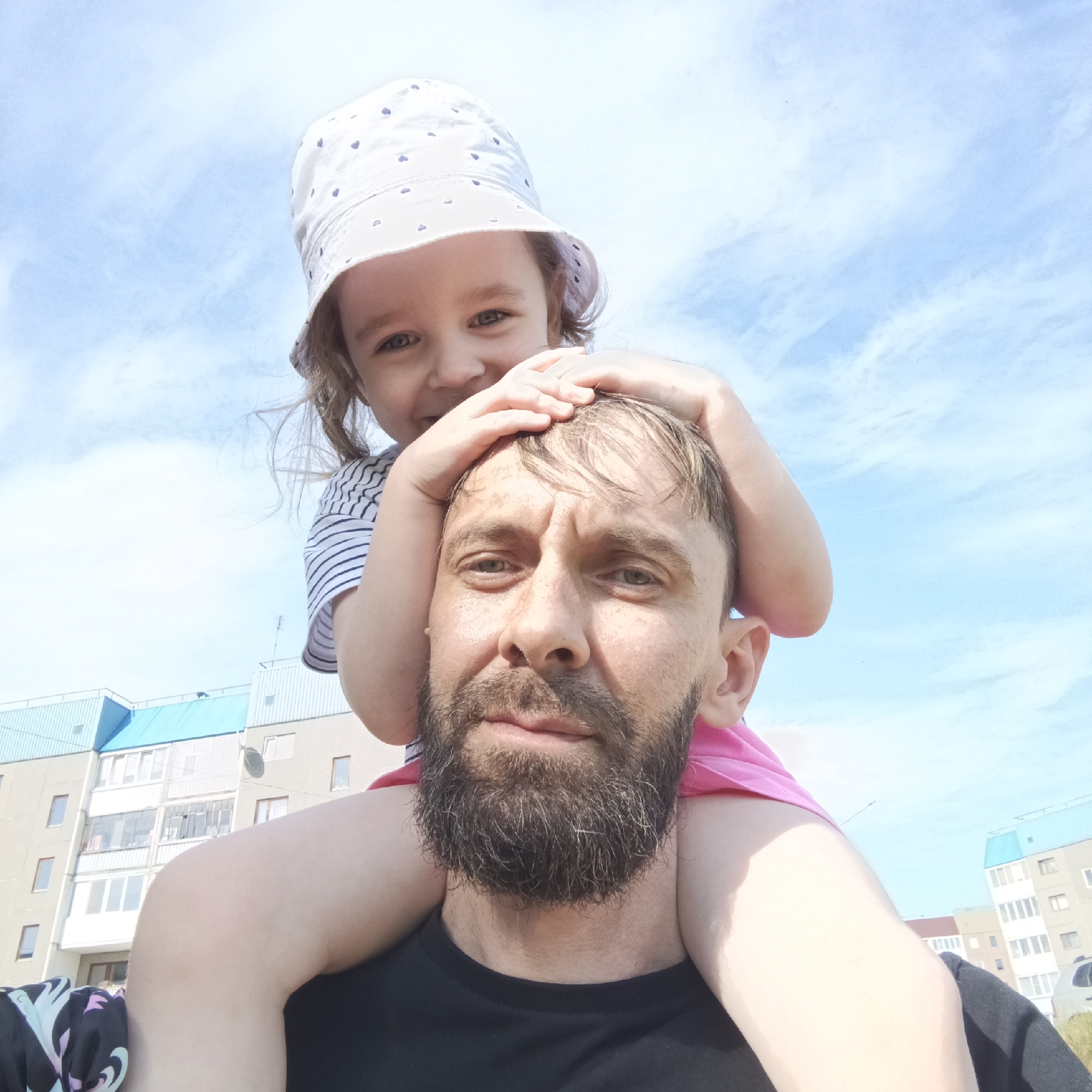A selfie captures a bearded man with a child on his shoulders against a bright sky with scattered clouds. The man, dressed in a black t-shirt, looks directly into the camera with a neutral expression. The child, wearing a white bucket hat with small blue heart patterns and a striped shirt, stands behind him, resting her hands on his head.
In the background, a multi-story building is visible, featuring a mix of gray and white exterior walls. The building has several windows and a blue roof. The image, potentially taken in Markovskii, Russia, emphasizes the relationship between the man and child in an outdoor urban setting.