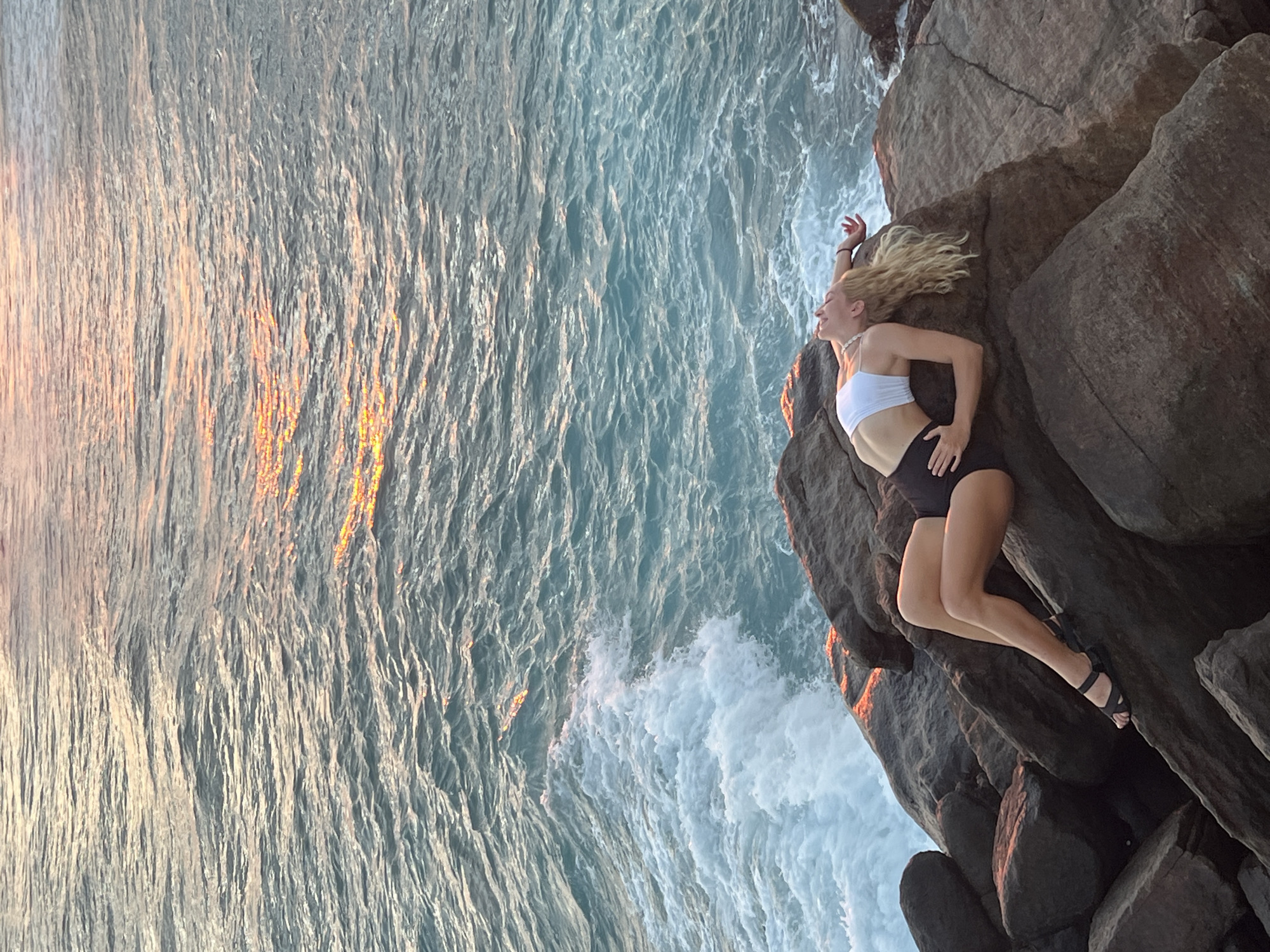 A woman in a white top and dark shorts lies on rocks near the water. Her blonde hair is spread out. She wears black sandals. The scene is Goviyapana, Sri Lanka. The water is rough with waves. Sunlight reflects on the water's surface.