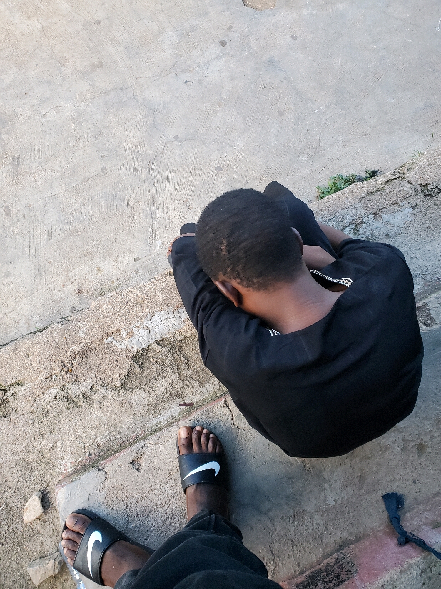 The image depicts a high-angle, overhead view of a young Black male sitting on concrete steps outdoors, likely in an urban environment. He is wearing a black t-shirt and black slide sandals with white Nike swoosh logos. His head is bowed, obscuring his face, and his arms are wrapped around his knees, suggesting he may be resting or contemplating. The concrete steps show signs of wear and tear, with cracks and variations in texture. To the left of the seated figure, another pair of legs wearing similar black slide sandals is visible, indicating the presence of another person, likely the photographer. The background consists of a concrete surface and some sparse greenery. There is no specific visual information within the image that would pinpoint the location to Jos, Nigeria; the scene appears to be a generic outdoor urban setting.
