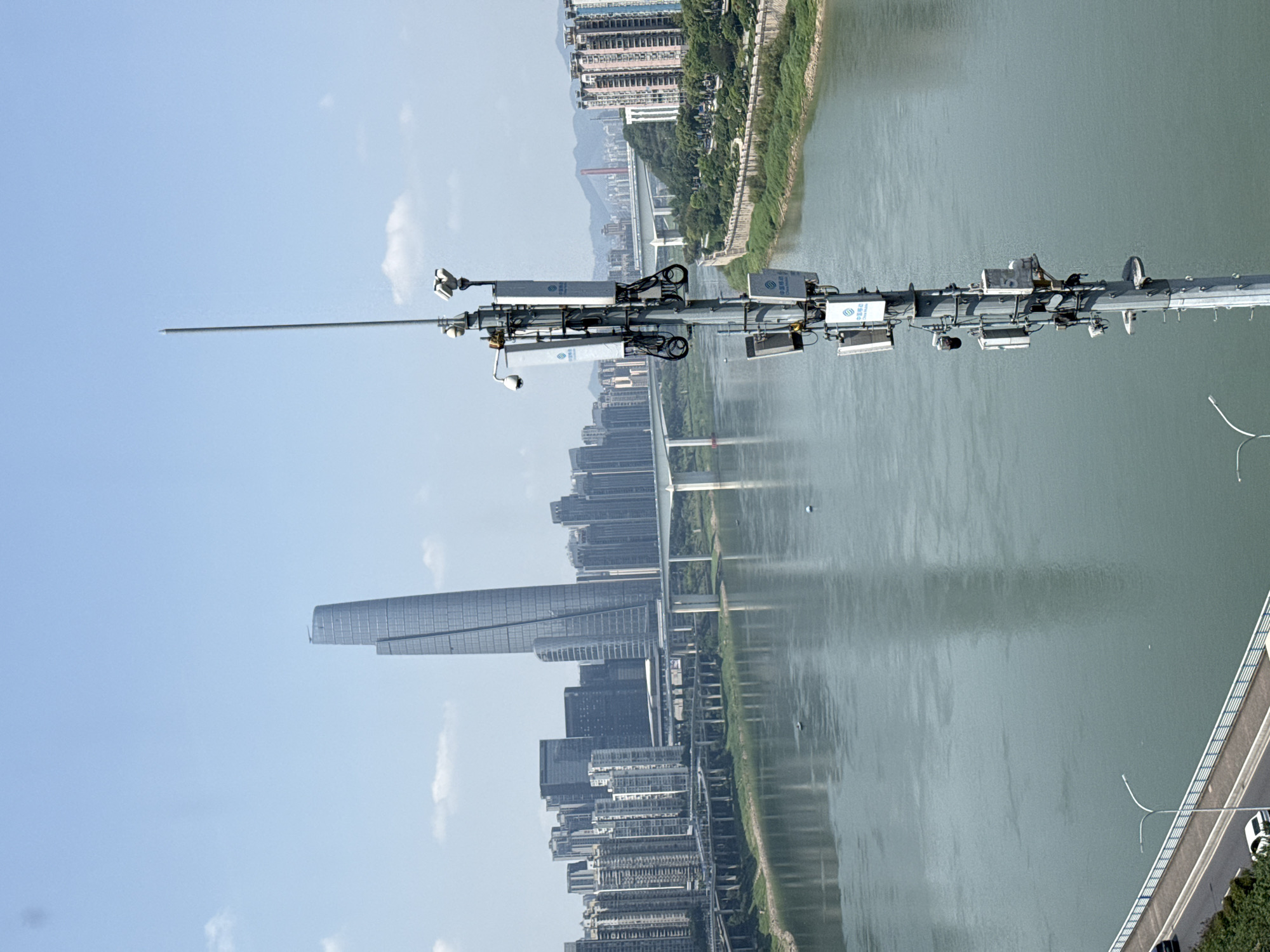 This image presents a vibrant urban landscape, likely taken from a bridge or high vantage point in Yuzhong District, China. The scene is dominated by a wide river flowing through a modern city under a clear blue sky with sparse white clouds.
In the mid-ground, a section of a bridge structure stretches horizontally across the frame, heavily equipped with various technological devices. Numerous white rectangular units, cameras, and intricate cabling are mounted on vertical poles attached to the bridge. A prominent, slender lightning rod or antenna extends vertically towards the sky from this assembly. On some of the white units, blue logos and the Chinese characters "中国移动" (China Mobile) are visible, suggesting telecommunications or monitoring equipment.
The wide river below the bridge reflects the bright sky and surrounding cityscape, displaying subtle ripples on its tranquil surface. On the opposite bank of the river, a dense and modern urban skyline rises, featuring numerous high-rise buildings. A particularly striking, tall, and slender skyscraper with a distinctive architectural design stands out against the backdrop of other residential and commercial towers. Multiple other bridges can be seen in the distance, spanning the river. The riverbanks are lined with some greenery and additional urban structures.
The time of day appears to be bright daytime, likely late morning or early afternoon, given the clear illumination. The weather is sunny and clear. No people or specific activities are visible, emphasizing the expansive infrastructure and modern development of the city.