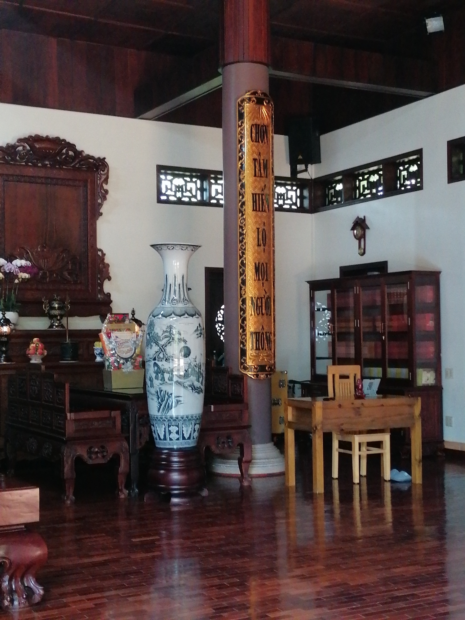 An indoor scene depicts a traditional room with dark wood furnishings and walls. A large, ornate wooden pillar stands in the foreground, adorned with a golden scroll featuring Vietnamese characters. To the left of the pillar, a tall, blue and white porcelain vase with intricate landscape designs sits on a dark wooden pedestal. Behind the vase, a dark wood cabinet with carved details is visible, topped with a smaller ornate wooden piece and a vase of flowers. A large, intricately carved wooden frame hangs on the wall to the left. To the right of the pillar, a wooden desk and a simple wooden chair are arranged. A dark wood cabinet with glass doors, filled with books and decorative items, stands behind the desk. A cuckoo clock is mounted on the wall above the cabinet. The floor is made of polished dark wood planks, reflecting the light from the room. The room appears to be a living or reception area, possibly in a home or a cultural establishment. The lighting is dim, with a soft glow illuminating the space. No people are visible in the image.