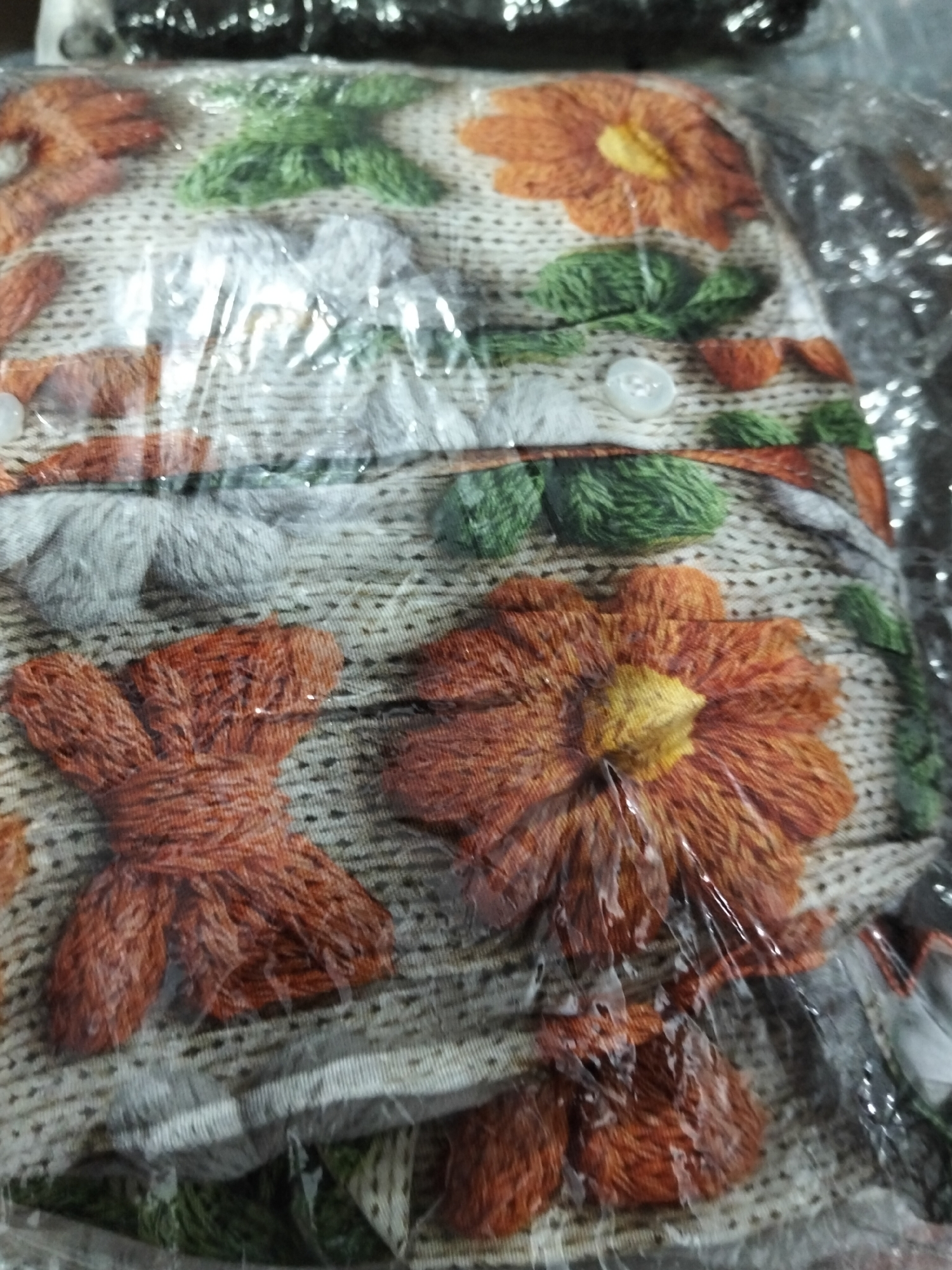 The image shows a close-up, top-down view of a fabric with embroidered floral designs and buttons, wrapped in clear plastic. The fabric is predominantly beige with embroidered elements in orange, yellow, green, and grey. Prominent orange flowers with yellow centers are visible, along with green leaves and stems. Some of the embroidery appears to be textured, resembling yarn. A few small, white buttons are also attached to the fabric. The plastic packaging creates reflections and distortions, obscuring some details. There are no people or recognizable geographical features of Payagaboh, Indonesia, in the image. The scene is static and appears to be a product intended for sale or storage.