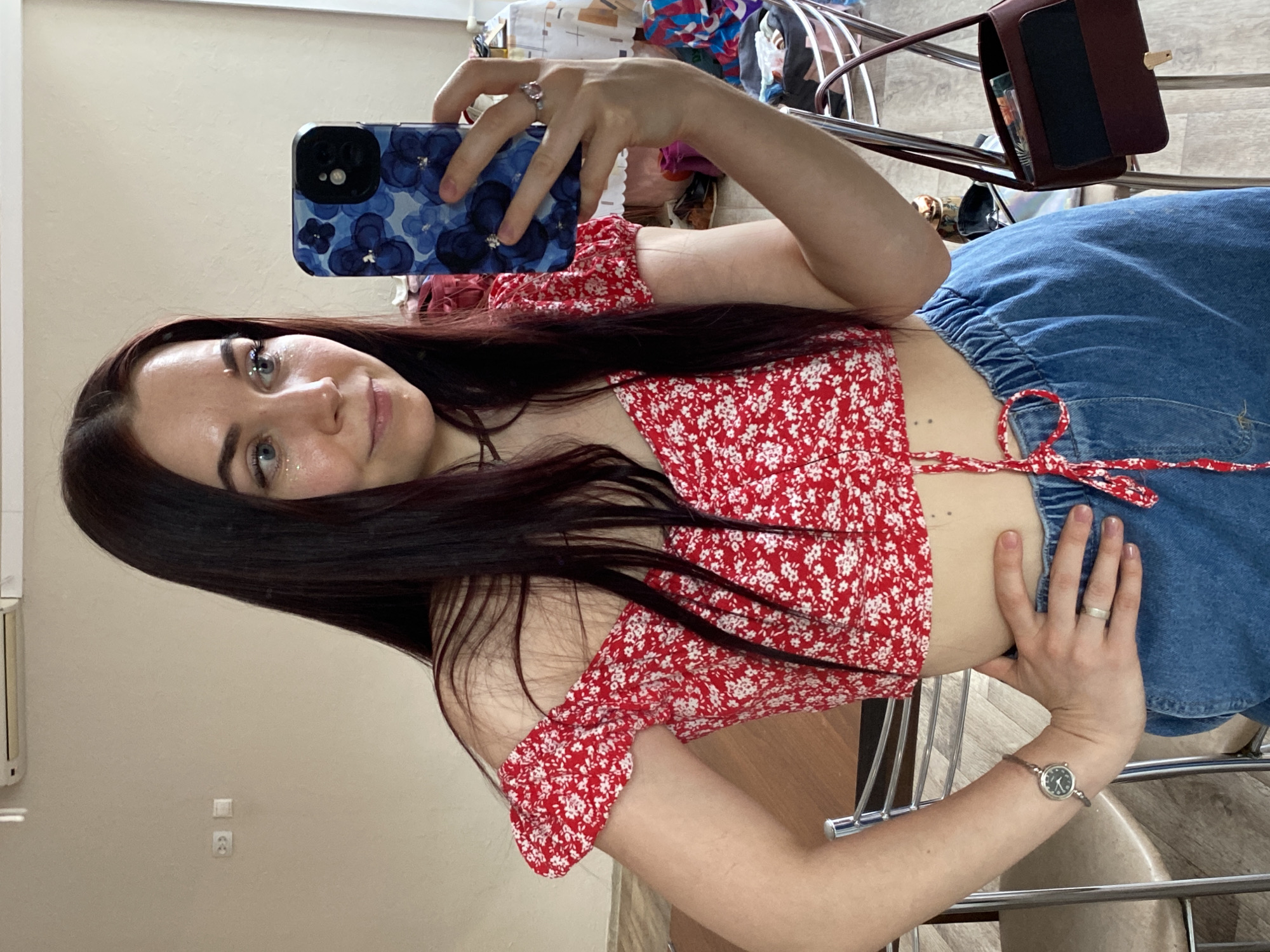 A woman is taking a selfie in an indoor setting. She is wearing a red floral crop top and blue denim bottoms. The phone in her hand has a blue floral case. She has long dark hair and appears to be wearing glittery makeup around her eyes. Her left hand has a ring on her finger. She's wearing a watch on her left wrist. There is a brown purse visible on a chair in the background, and a mess of items behind the chair. The location appears to be a room in a building, potentially in the city of Chaikovsky, Russia. The walls are a neutral color.