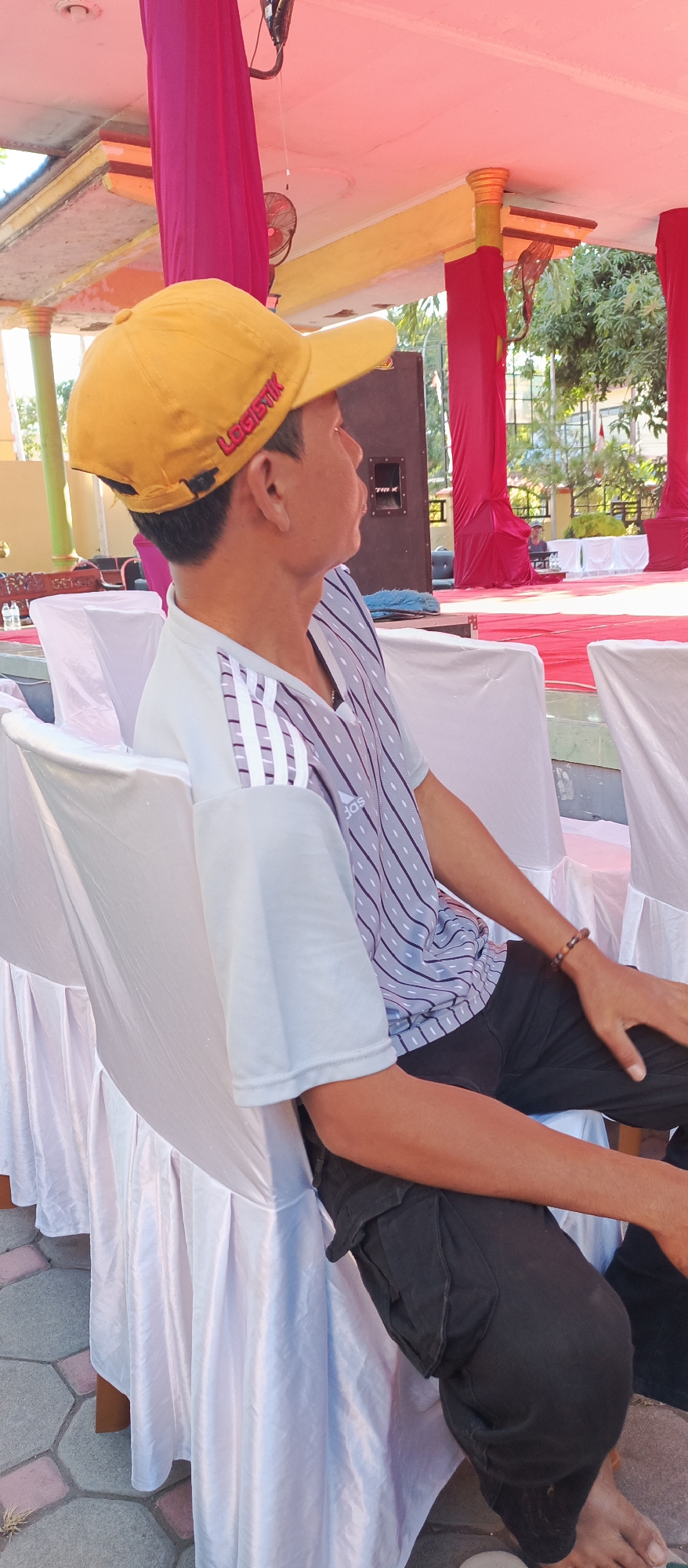 In Arjasa, Indonesia, a man wearing a yellow cap with the word "LOGISTIK" and a striped t-shirt sits on a white-covered chair. He is looking to the right, with his left arm resting on his lap and his right arm extended, holding onto the chair. The background features red drapes, a speaker, and a paved ground.
