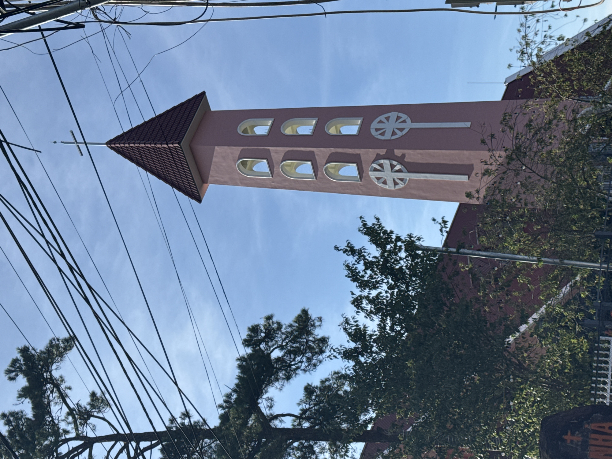 The image shows a pink church tower with arched windows and cross emblems, located in Đa Lộc, Vietnam. The tower is topped with a red, tiled, pyramid-shaped roof and a cross. The surrounding environment includes trees with green foliage, power lines crisscrossing the sky, and a blue sky with some clouds. The lower portion of the image shows part of the church building with a red roof and some fencing. There are no people visible in the image.