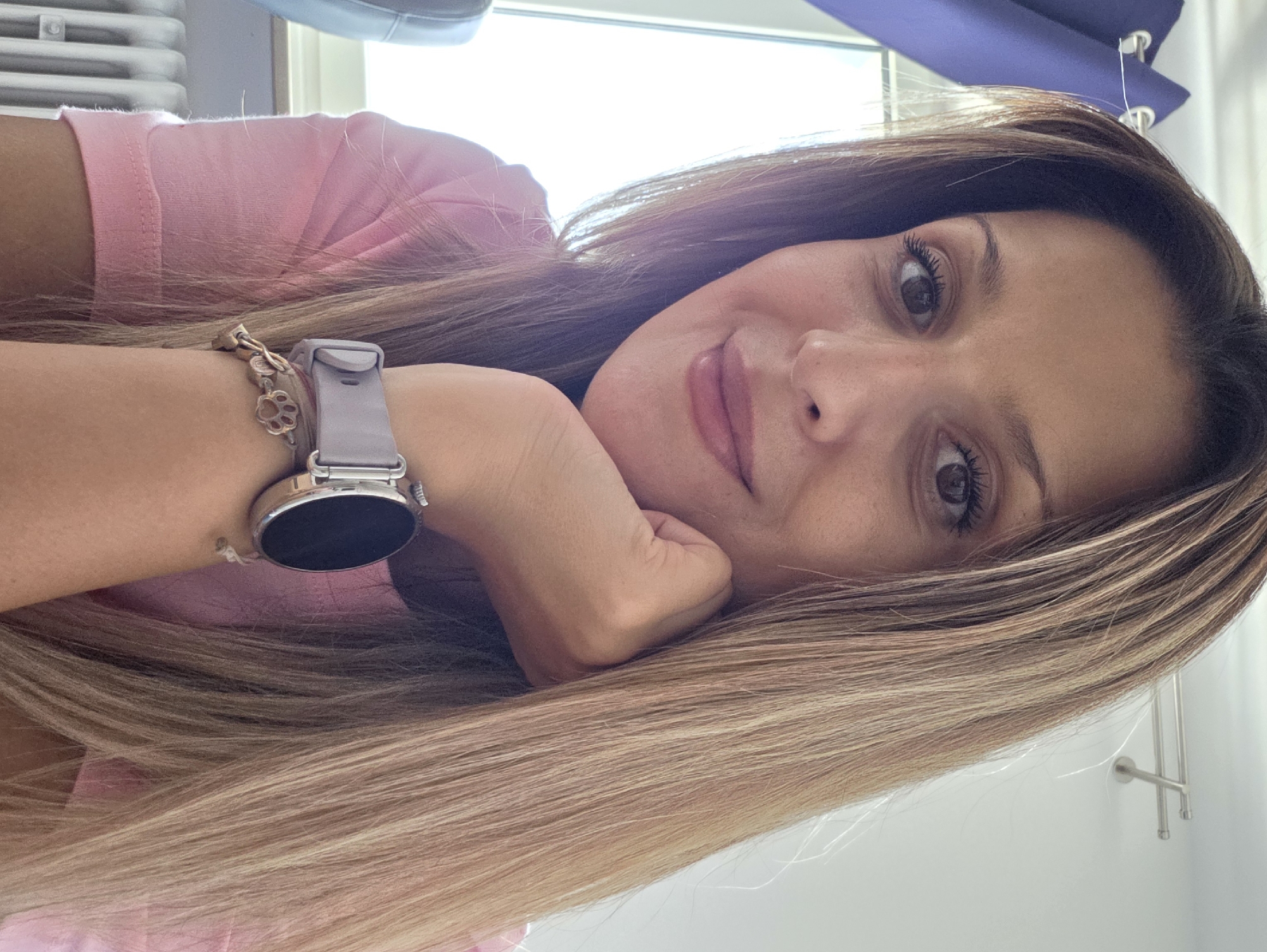 A woman is resting her chin on her hand, looking at the camera. She has long, blonde hair and is wearing a pink t-shirt. She has a smartwatch on her left wrist with a light pink band and a silver watch face. There are some charms on the band of the watch. She is indoors, with a window and blue curtains visible in the background, and a radiator below the window. The scene is likely an indoor residential or office setting in Munich, Germany.