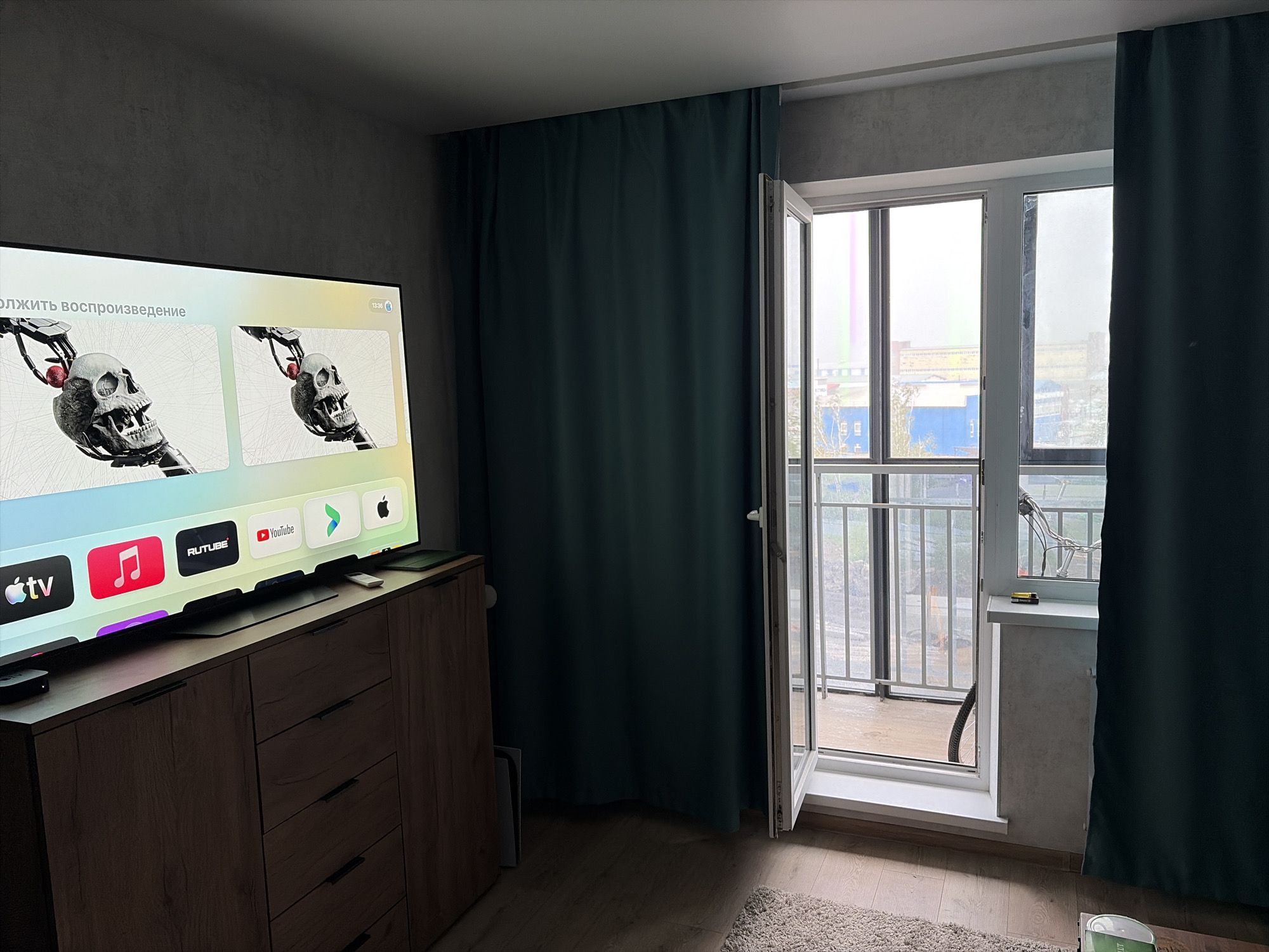 The image depicts a modern, indoor living space, likely an apartment, furnished with a large television and a wooden cabinet. The television screen displays an interface with various application icons, including Apple TV, a music note, Rutube, and YouTube, suggesting it is a smart TV or connected to a media player. A dark teal curtain is visible, partially drawn across a large window or balcony door that offers a view of an urban industrial landscape. The exterior view includes buildings and a railing, indicative of a balcony or external walkway. The room is lit by natural light coming through the open window, casting shadows on the wooden floor. The overall atmosphere is neat and contemporary. The specific location of Yekaterinburg, Russia, cannot be determined from visual cues alone; this information is extraneous to the image content.