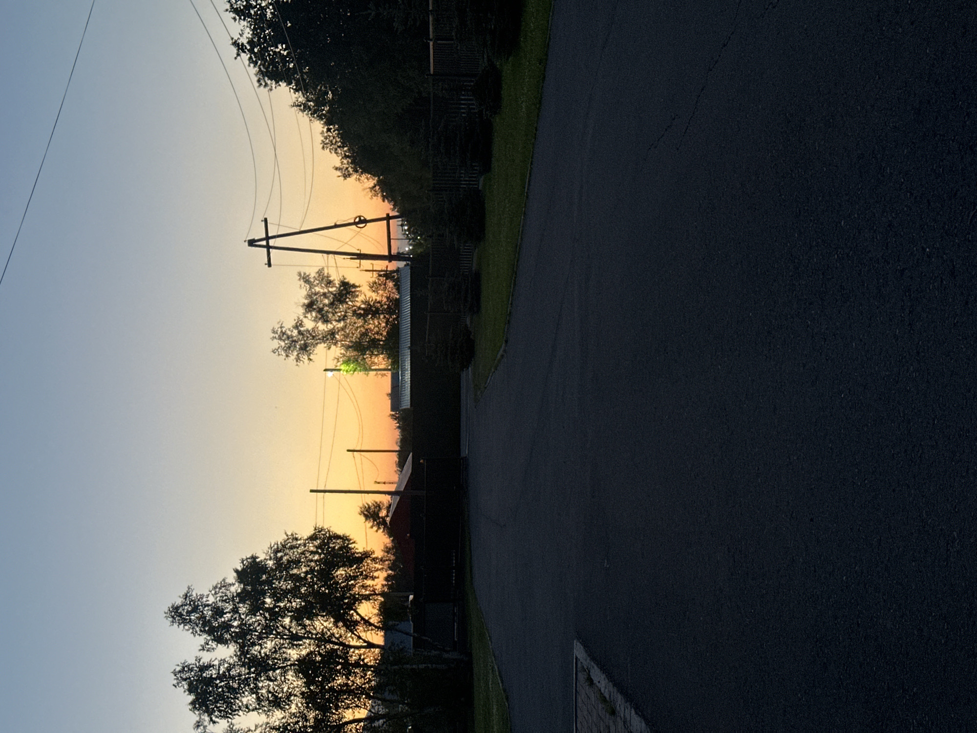 A paved road curves through a rural landscape at dusk. On the left, silhouetted trees line a dark fence. Beyond the fence, indistinct buildings and more trees are visible against a sky transitioning from pale blue to orange. Utility poles with wires stretch across the scene. On the right, a strip of lawn dotted with small, dark evergreen trees runs along the fence. The road itself is dark and textured, with a small paved area on the left edge. The overall impression is of a quiet evening in a sparsely populated area.