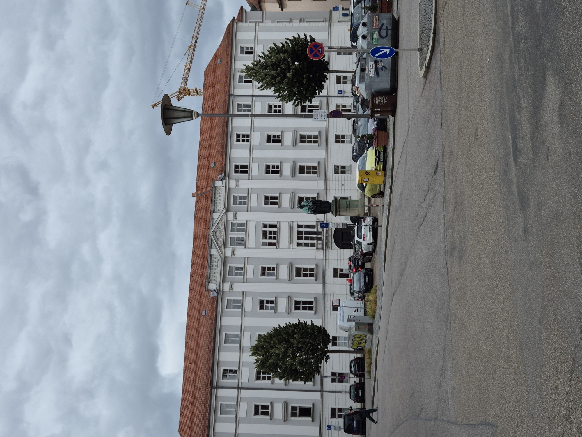 A building with a red tiled roof and a gray facade with white vertical stripes dominates the scene. The building has numerous windows and an arched entrance. In front of the building, a parking lot is visible with several cars and a statue of a person in a green robe. A person is walking in the foreground. There are trees, streetlights, and a construction crane in the background. The scene appears to be located in Regensburg, Germany, near the Donau-Einkaufszentrum.