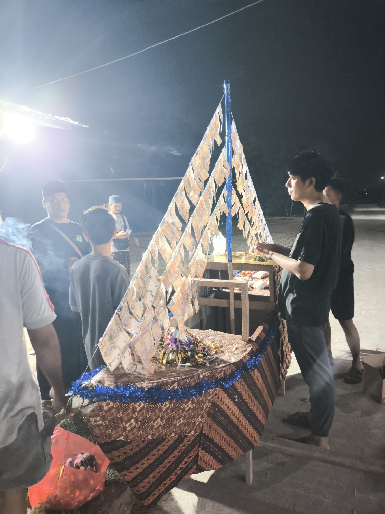 In the city of Cilegon, Indonesia, at night, a group of people are gathered around a decorated structure. The structure is shaped like a boat, with a tall, triangular mast adorned with numerous Indonesian Rupiah banknotes. The boat itself is covered with a patterned fabric and decorated with blue tinsel. A table is placed on top of the boat, holding some items that are not clearly discernible. Several individuals, appearing to be young men and older men, are standing and observing the structure. Some are looking at the banknotes, while others are engaged in conversation. The scene is illuminated by artificial lights, creating a somewhat dim and hazy atmosphere. There is also a pink plastic bag on the ground containing what appear to be items wrapped in foil, possibly food or offerings. The ground is a paved surface, and in the background, there are hints of buildings and outdoor lighting.