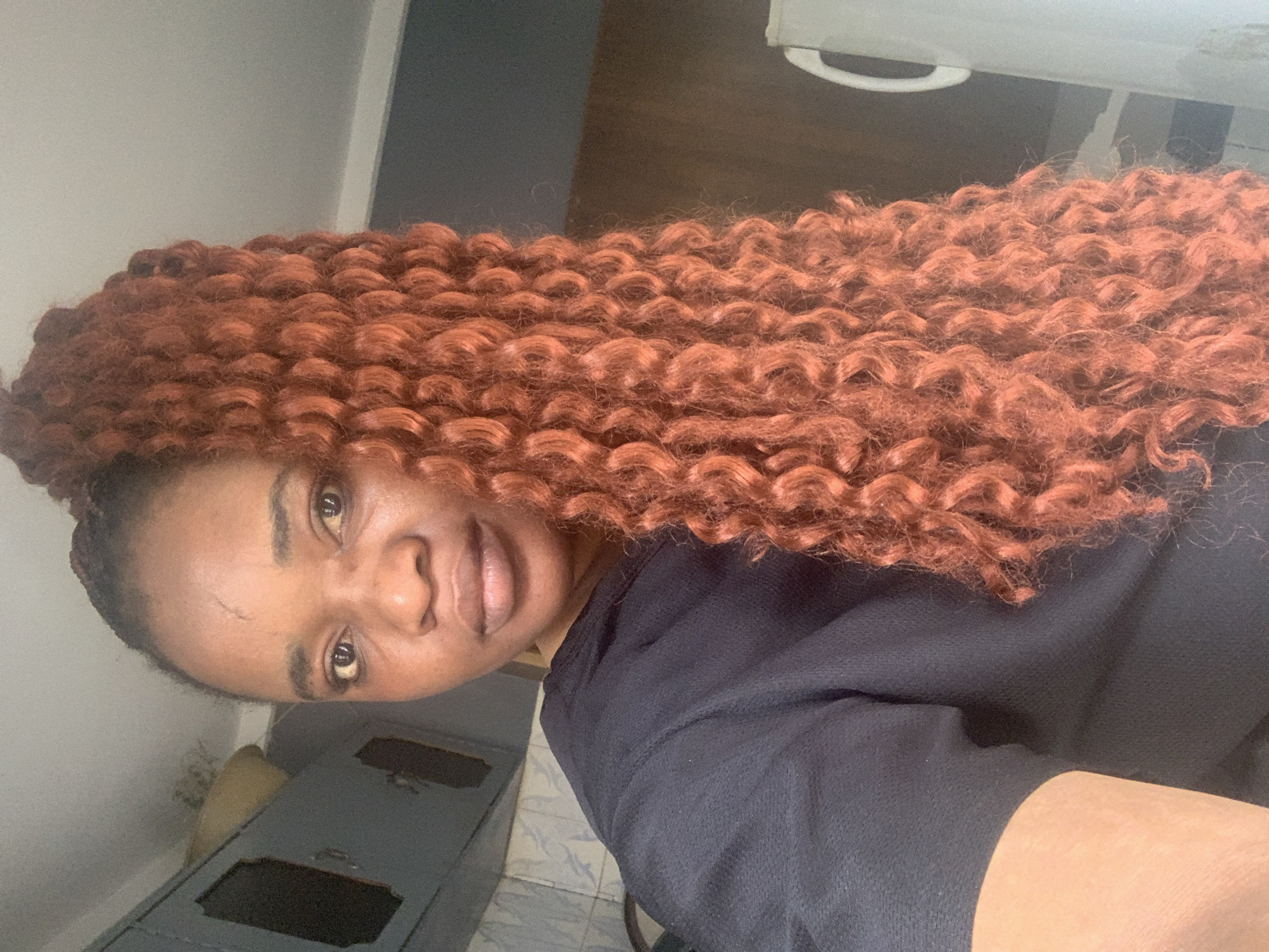 A medium close-up, portrait-oriented selfie features a woman with brown skin and long, voluminous reddish-brown or auburn curly hair. The curls appear to be a protective style, possibly crochet braids, with a consistent, tight coil pattern. Her hair cascades over her right shoulder and chest.
The woman is looking directly at the camera with a neutral expression and a slight, closed-mouth smile. A distinct scar or mark is visible on her forehead, above her left eye. She has dark, neatly shaped eyebrows. She is wearing a plain black short-sleeved top.
The background suggests an indoor setting. To her left, a light-colored wall is visible, along with a piece of dark-colored wooden furniture that appears to be a cabinet or chest of drawers with decorative cut-outs and metallic handles. Above her head, a darker wall or surface transitions into a lighter area, with a white, curved object that could be a handle or part of an appliance. The lighting is soft and natural, indicating daytime. There is no visible text in the image. The image is taken in a residential interior in Jos, Nigeria.
