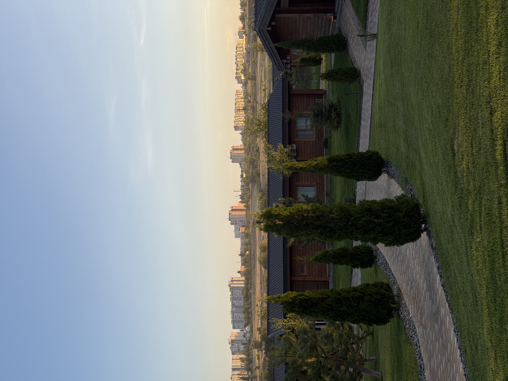 The image depicts a modern resort or residential complex in the city of Kamyshin, Russia, during the early morning or late evening, as indicated by the soft, golden light. In the foreground, manicured green lawns are interspersed with winding pathways made of light-colored paving stones. Several evergreen trees, likely cypresses or similar varieties, are strategically placed along the paths, adding a touch of natural beauty.
Behind the landscaped grounds, a row of rustic wooden cabins or houses is visible. These structures feature dark-tiled roofs and natural wood exteriors, suggesting a contemporary architectural style that blends with the environment. Some cabins have visible windows and air conditioning units.
In the background, a sprawling cityscape rises against the horizon under a clear, pale blue sky. Numerous apartment buildings of varying heights and architectural styles dominate the skyline, with some appearing older and others more modern. The distant landscape is characterized by rolling hills and sparse vegetation. The overall scene conveys a sense of peacefulness and well-maintained modernity juxtaposed with the urban sprawl in the distance. There are no people or animals visible in the image.