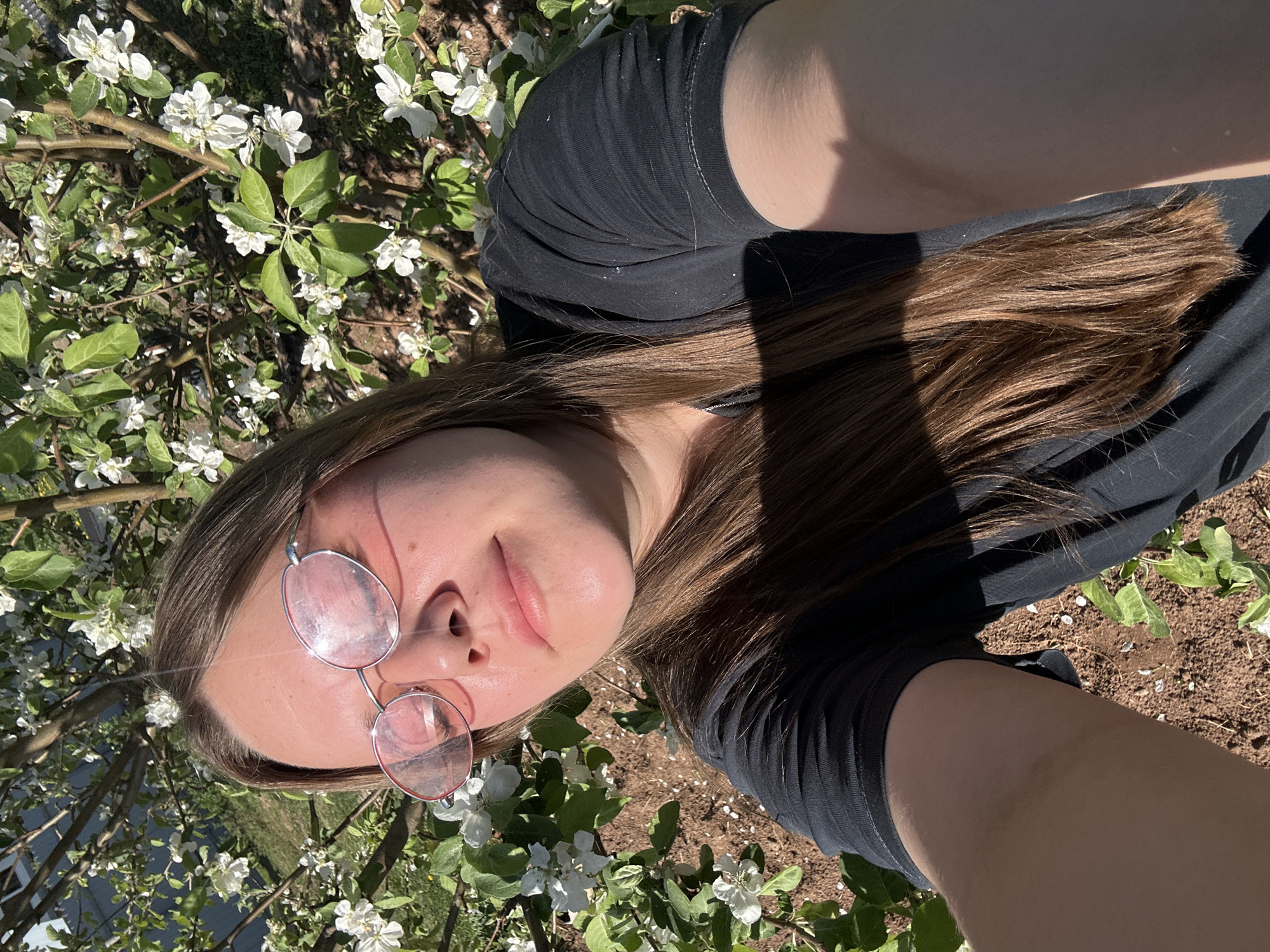 A selfie shows a woman with light brown hair wearing glasses and a dark t-shirt, positioned among flowering branches. The backdrop includes green foliage and white blossoms. The ground appears to be covered in reddish-brown soil or mulch. The location is supposedly in Ольховка, Russia, based on prompt information. The woman appears to be outdoors in daylight.