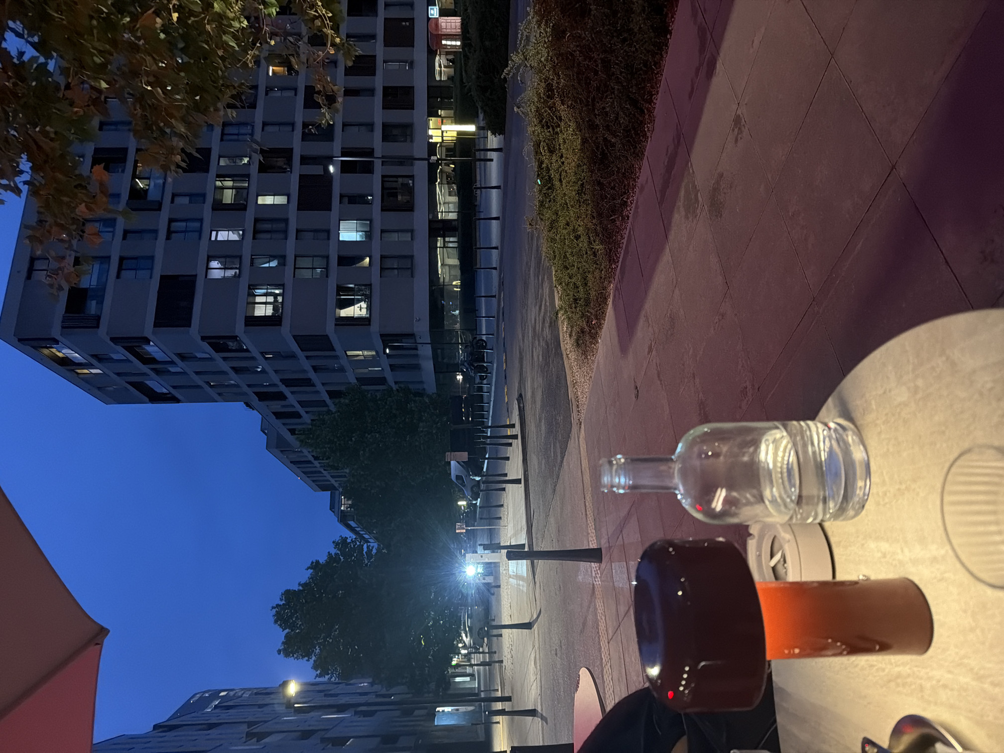 The image depicts an outdoor scene at dusk, likely in a modern urban setting. In the foreground, a table with a reddish-brown lamp and a clear glass bottle filled with water is visible. Beyond the table, a sidewalk with large square tiles leads to a street. The street is lined with bollards and appears to be a public space with parked cars and a street light. In the background, several contemporary buildings with many windows dominate the skyline. Trees are present, some with autumnal foliage. The sky is a deep blue, indicating the transition from day to night. The lighting suggests artificial illumination from streetlights and building interiors. There are no discernible people actively engaged in any actions, though figures might be implied within the illuminated windows of the buildings. The environment appears to be a pedestrian area adjacent to a street, likely a part of a city designed with modern architecture.