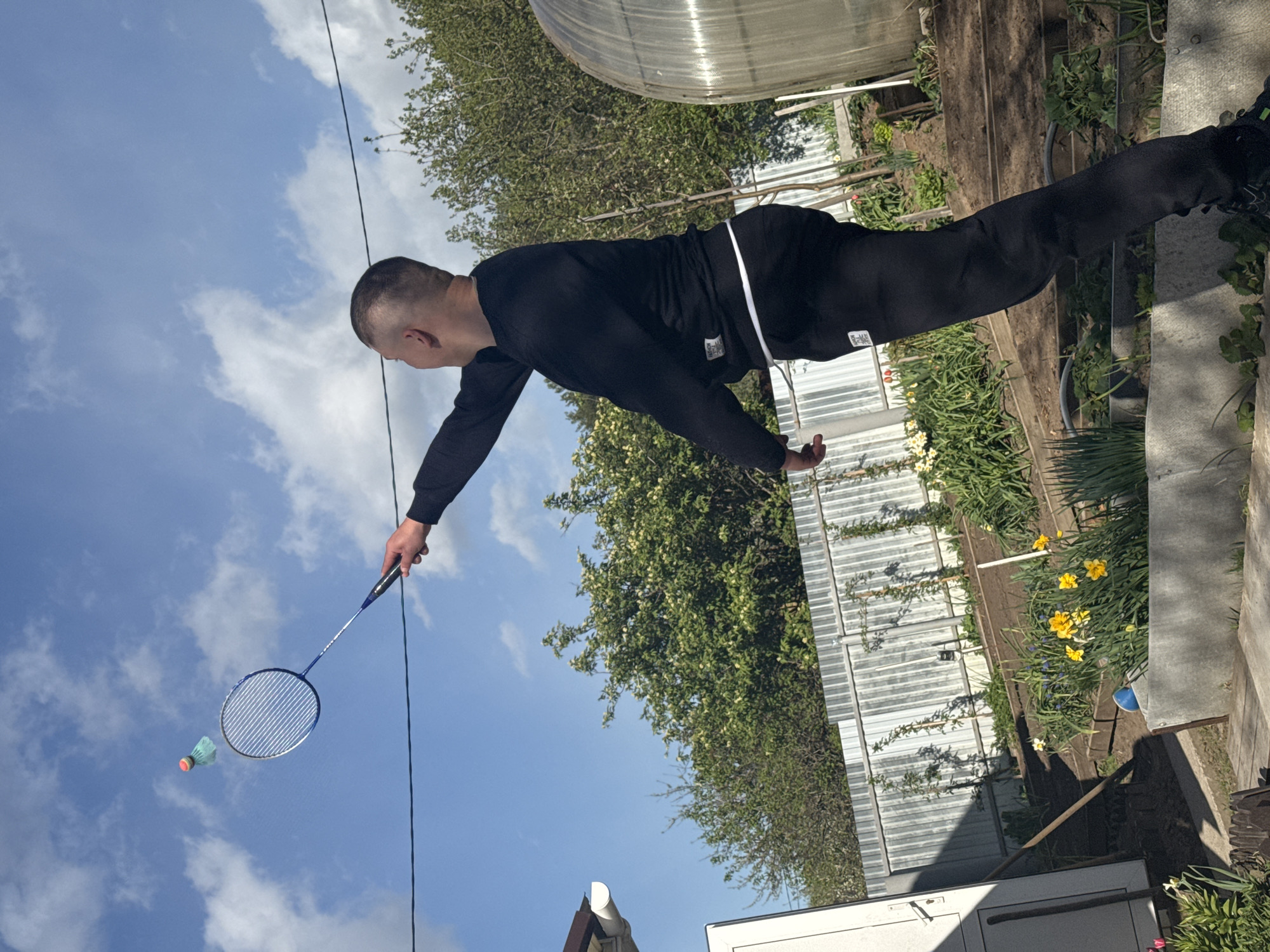 A man is playing badminton in an outdoor setting. He wears a black long-sleeved shirt and black pants, and is holding a blue badminton racket. A blue and green shuttlecock is in the air, presumably just hit by the racket. The background includes a blue sky with white clouds, green trees, and a grey fence. The location is the city of Chaikovsky, Russia. There is a building visible in the background. Flowers are blooming in a garden bed next to the fence. A greenhouse structure can be seen as well. A utility pole or wire runs across the sky.
