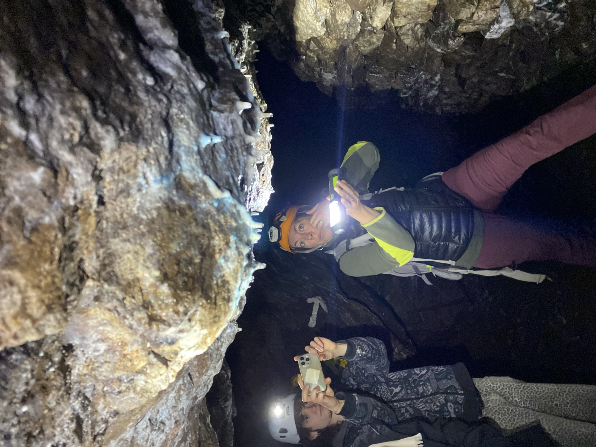 This media file captures two individuals engaged in cave exploration within a dark cave environment in Les Chavannes-en-Maurienne, France. The scene is predominantly dark, illuminated by artificial lights from headlamps and a flashlight, revealing rugged rock formations with earthy tones and some glistening mineral deposits, including small, pointed stalactites or similar formations on the cave walls, some appearing pale blue or white.
The individual positioned towards the top of the frame wears an orange helmet equipped with a headlamp, a grey and lime-green long-sleeved top, and a dark puffer vest. They hold a bright flashlight with their right hand, directing its beam upwards, and look directly at the camera with a wide-eyed, possibly surprised or excited expression. A ring is visible on their left hand. Their lower body is clad in maroon-colored pants.
The second individual, positioned towards the bottom, wears a white helmet with a "Petzl" brand headlamp and a dark, patterned jacket. They are actively using a light-colored smartphone, held horizontally with both hands, to capture a photo or video, with their face partially obscured by the device.
A notable detail on the dark cave wall between the two individuals is a distinct white symbol resembling a pickaxe or hammer, likely serving as a trail or directional marker. The image effectively portrays the challenges and visual interest of caving, with specialized gear being essential in the dimly lit, natural underground setting.