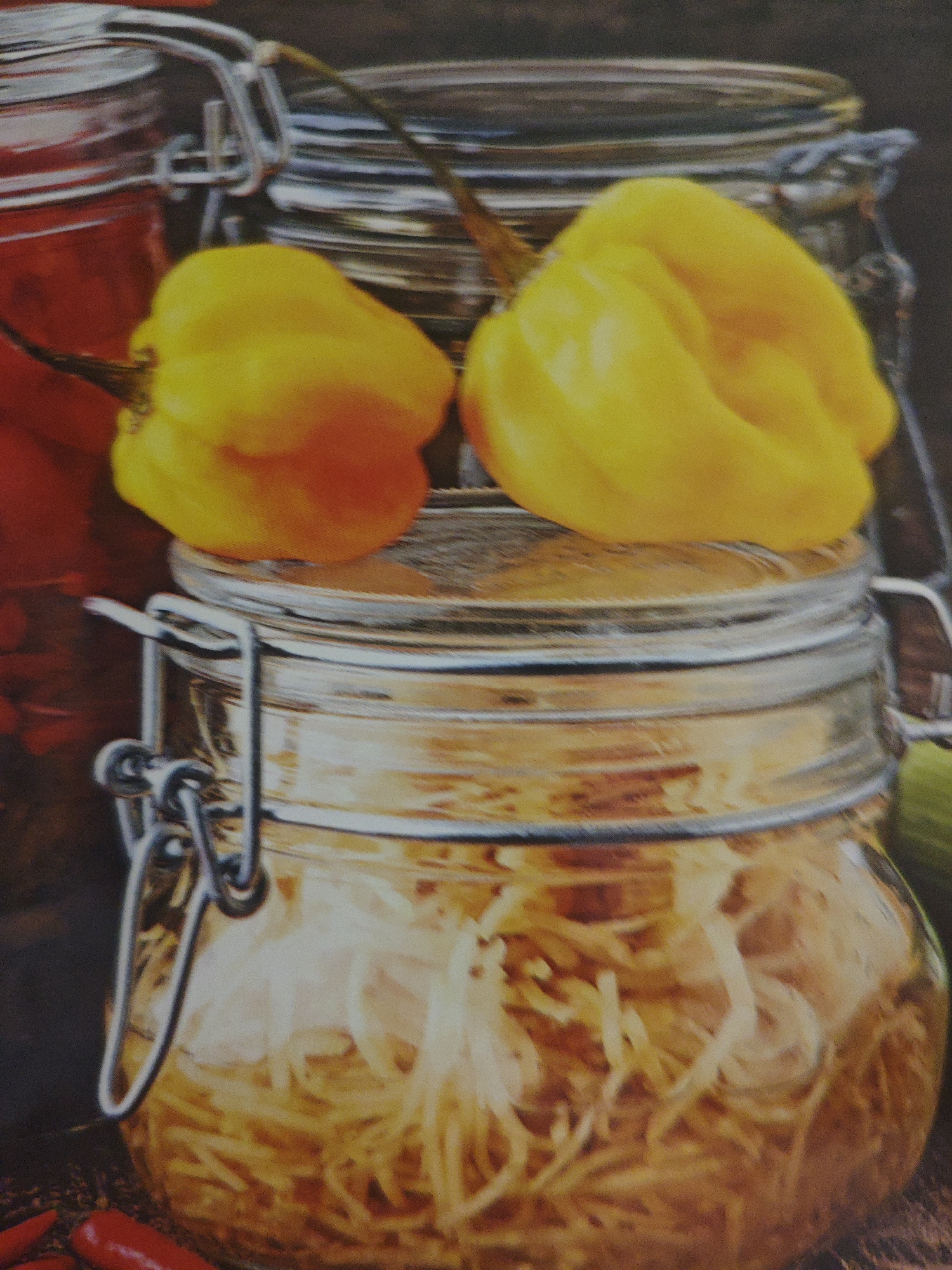 This is a close-up, overhead shot of two glass jars with clamp lids, one stacked on top of the other. The top jar contains two bright yellow habanero peppers resting on the rim. The bottom jar is filled with thinly sliced, translucent ingredients, possibly pickled or fermented vegetables, submerged in a golden liquid. The jars are on a dark, textured surface, possibly a wooden table. Two red chili peppers are partially visible in the bottom left corner. The image appears to be a still life, likely for a culinary or food-related context, and there is no indication of its location in Armavir, Russia.