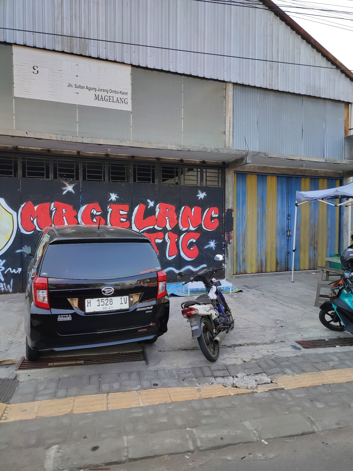 The image depicts a street scene in Magelang, Indonesia, in front of a building with a corrugated metal facade. A white sign with black text reads "Jln. Sultan Agung Jurangombo-Karet MAGELANG". A black Daihatsu Sigra car with license plate H 1528 IV is parked on the street. A white and blue motorcycle is parked behind the car, and a teal scooter is parked to the right. Graffiti covers the wall behind the car, featuring red lettering that reads "MAGELANG T". The building has a section with a blue and yellow striped roll-up door. A white canopy tent is set up to the right of the building. The street is paved with concrete blocks and has a yellow tactile paving strip. The overall scene suggests a commercial or industrial area.