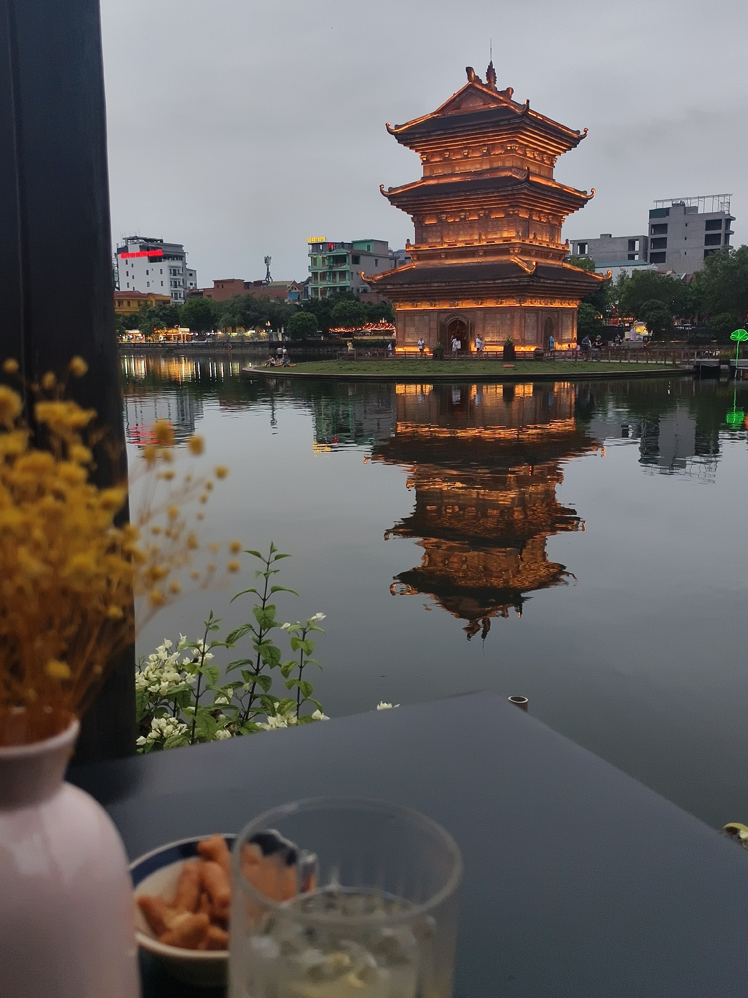 The image depicts a serene twilight scene at a lake, likely in Hoa Lư Ward, Vietnam, with a prominent pagoda as the central subject. The pagoda, illuminated by warm, golden lights, stands tall with multiple tiered roofs and intricate architectural details. Its reflection is clearly visible in the calm water, creating a symmetrical and captivating visual.
In the foreground, a blurry view of a table is visible, with a glass of a light-colored beverage and a small bowl containing snacks. A light pink vase and dried yellow flowers are also in the foreground, adding a touch of color and texture to the composition, though they are out of focus. Behind the pagoda, several modern buildings and trees line the shore, suggesting an urban or semi-urban environment. The sky is overcast, indicating either dawn or dusk.
The atmosphere is peaceful and tranquil. A few people can be seen near the pagoda and along the water's edge, appearing to be enjoying the evening ambiance. The overall mood is one of quiet reflection and appreciation for the scenic beauty of the location. The time of day is likely dusk, given the artificial lighting and the soft, diffused sky.