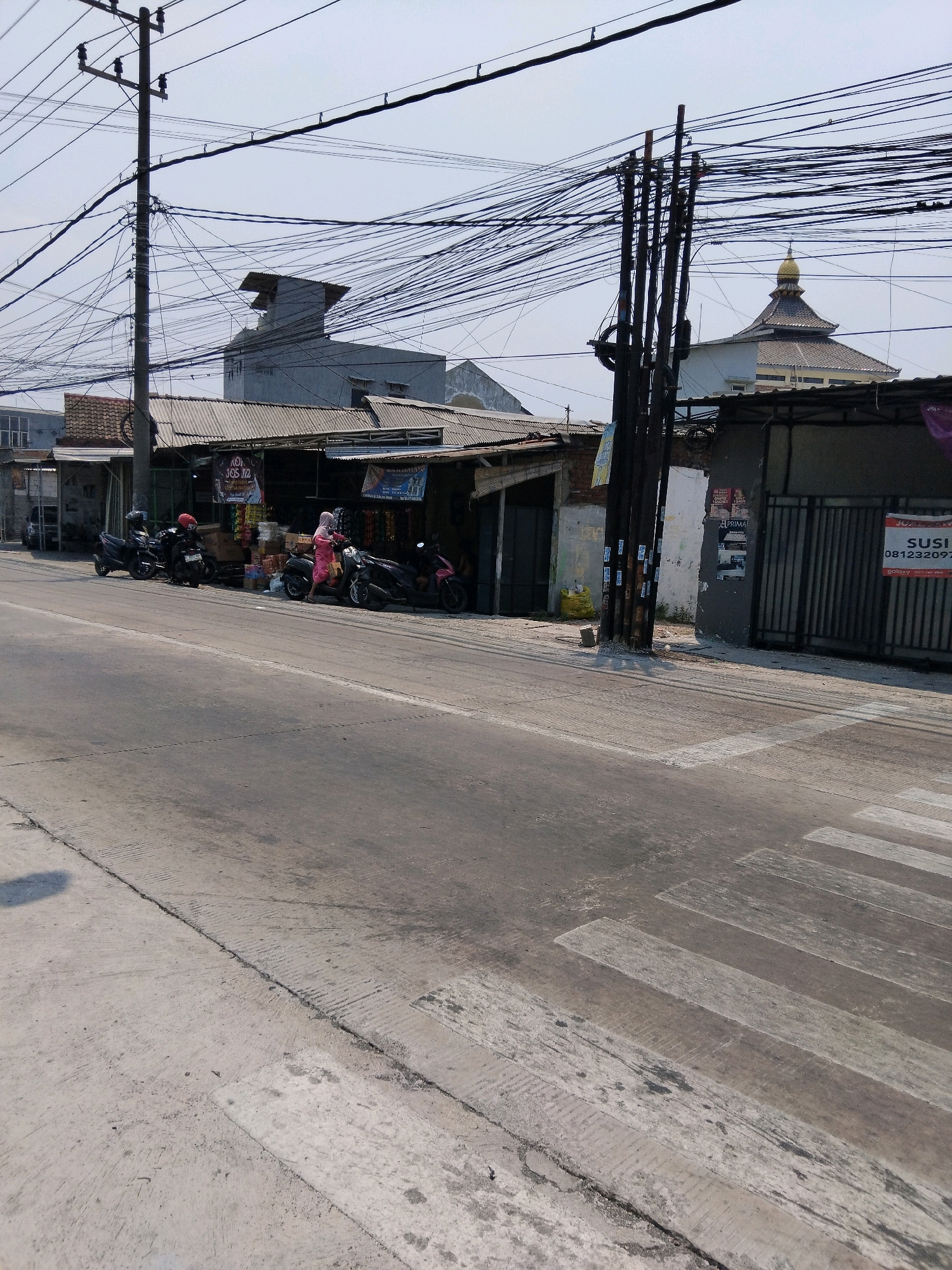 The image depicts a street scene in the city of Kepuh Kiriman, Indonesia, characterized by a dense network of overhead utility wires. In the foreground, a paved road with a visible pedestrian crossing runs horizontally across the frame. To the right of the road, a row of small shops with corrugated metal roofs are situated. Several motorcycles are parked in front of these shops, and two individuals are standing near them. One person, wearing a pink headscarf and a pink garment, appears to be interacting with a motorcycle. Further back, a large building with a dome, likely a mosque, is visible above the shops. A tall utility pole with numerous cables is prominent on the left side of the image, contributing to the cluttered overhead environment. The sky is overcast.