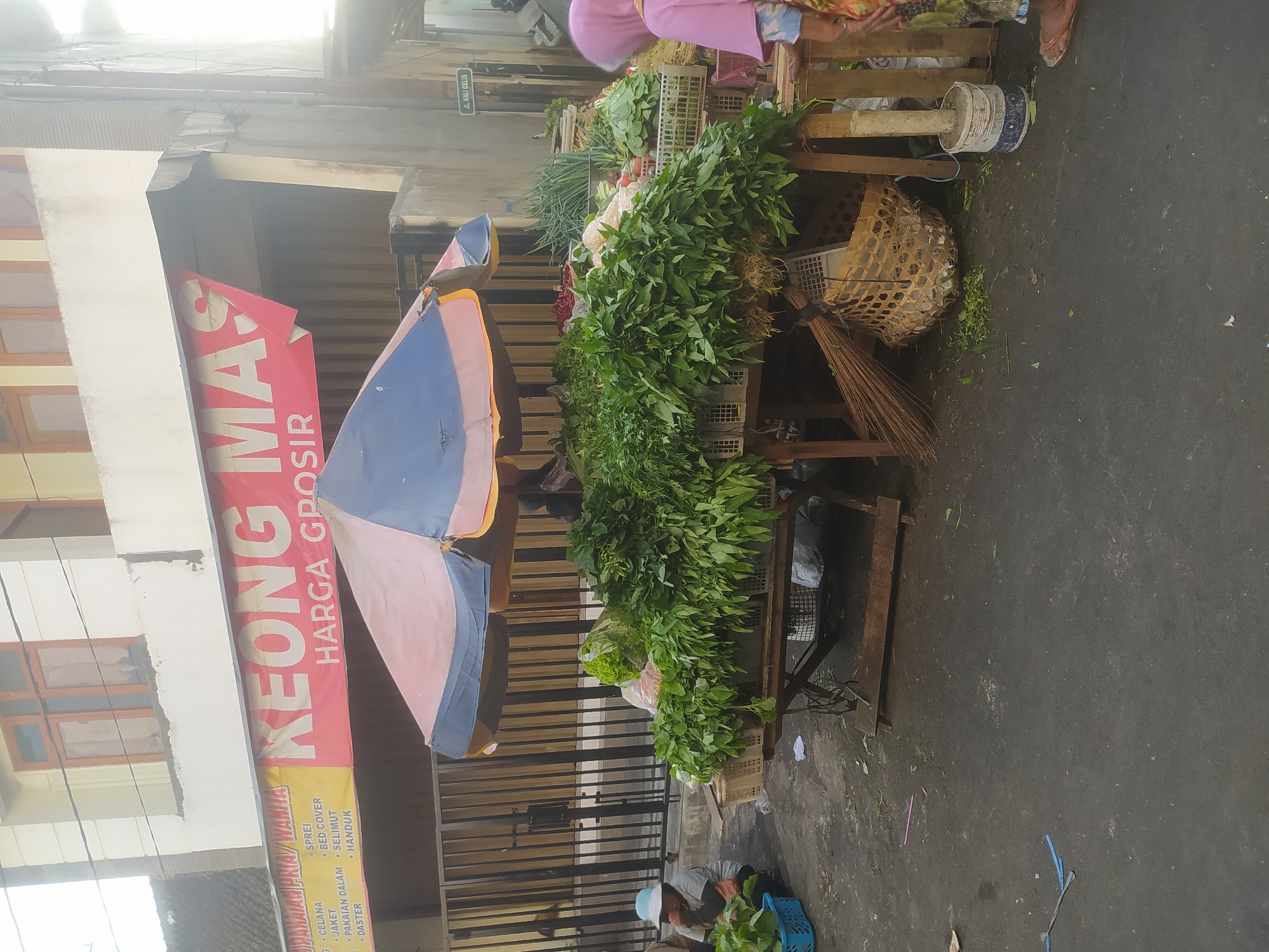This image depicts an outdoor vegetable stall in Salatiga, Indonesia. A banner overhead reads "KEONG MAS HARGA GROSIR," indicating the name of the establishment and that it sells wholesale. The stall is covered by a colorful umbrella, and displays a variety of fresh vegetables, including leafy greens and what appear to be scallions. A person in a pink hijab stands to the right, seemingly interacting with the stall. To the left, a person wearing a light blue hat is crouched down, examining some vegetables in a blue basket. The ground is paved and appears to be a street or market area. In the background, a building with windows is visible. A street sign that reads "JL. KALI GELIS" is mounted on a pole to the right of the stall. The overall scene captures a moment of commerce at a local market.