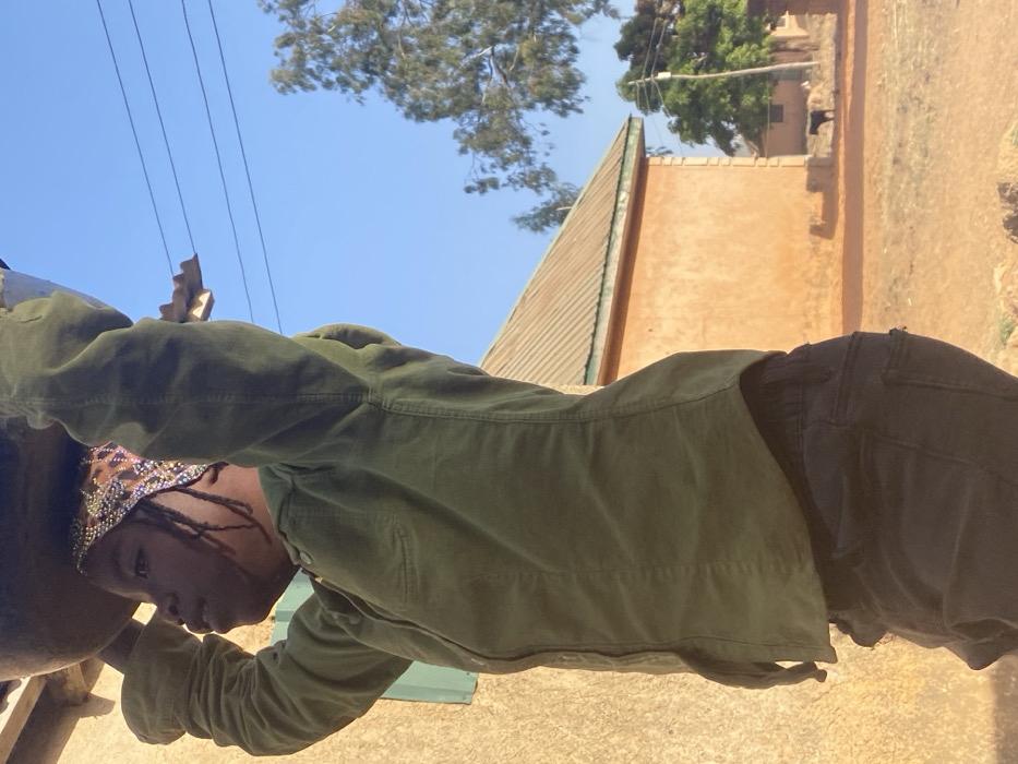An outdoor scene in Udu, Nigeria, depicts a young individual in profile, facing left, carrying a large, dark basin on their head, hands raised for support. The individual wears an olive green long-sleeved collared shirt, dark trousers, and a patterned headwrap over braided hair. The immediate left foreground shows a light brown building wall with a partially visible green shutter. In the midground, under a clear blue sky with overhead utility wires, an orange-walled building with a corrugated metal roof stands beside a dusty, reddish-brown unpaved area. Further back, additional buildings and trees with sparse foliage are visible. A dark-colored animal, consistent with a dog, walks in the distant background on the unpaved ground. Sunlight illuminates the scene, indicating daytime conditions.