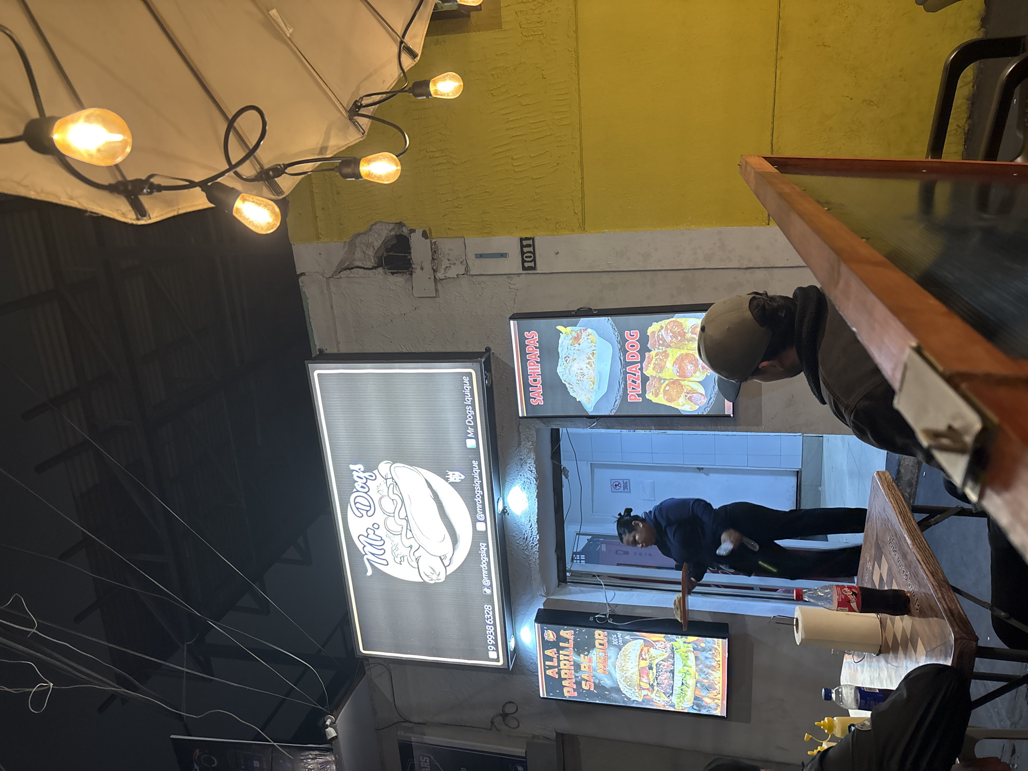 This image depicts an outdoor dining area at night, likely a food stall or restaurant, located in Iquique, Chile. The focal point is a brightly lit sign for "Mr. Dogs," featuring a stylized hot dog illustration. Below this, there are additional illuminated signs advertising menu items such as "A LA PARRILLA SABE MEJOR" (Grilled tastes better) with a burger image, and "SALCHIPAPAS" (sausage and fries) with "PIZZA DOG" below it.
A woman, appearing to be an employee, is visible near the entrance, holding a tray with food. She is wearing a dark blue uniform. In the foreground, a table is set with a bottle of Coca-Cola, a roll of paper towels, and condiments. Two individuals are seated at the table, their backs partially to the camera. One person is wearing a dark jacket and baseball cap. The environment is illuminated by string lights, some of which are visible hanging from a canopy. The building facade is textured and painted in yellow and gray. A building number, "1011", is visible on the wall. The overall scene suggests a casual, late-night dining establishment.