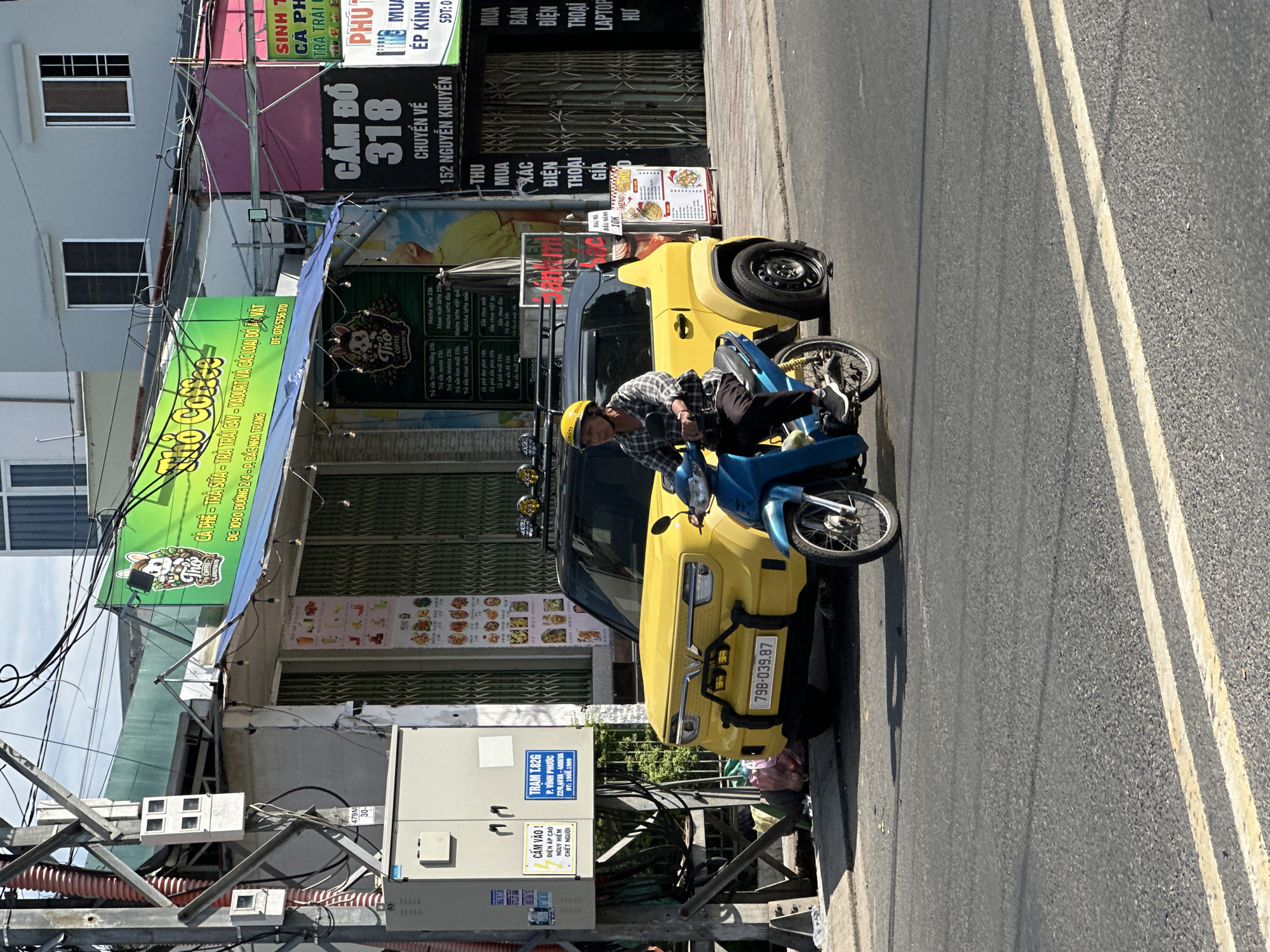 This image captures a vibrant daytime street scene in an urban area of Nha Trang, Vietnam, bathed in bright sunlight. The main subjects are a person on a blue scooter and a yellow compact car, both parked on the side of a road marked with double yellow lines.
The person, wearing a plaid shirt and a yellow helmet, is seated on the blue scooter, facing left, with a relaxed posture. The bright yellow car, possibly a compact SUV or crossover, is parked directly behind the scooter, featuring a front bumper guard and a visible license plate that reads "798-039.87."
Behind the vehicles, several shopfronts line the street. A prominent green awning advertises "CÀ PHÊ - TRÀ SỮA - TRÀ TRÁI CÂY - YAGOURT VÀ CÁC LOẠI ĐỒ ĐÁT COFFEE" (Coffee - Milk tea - Fruit tea - Yogurt and various ice cream), with a phone number "0765756770" and the address "ĐC: 1090 ĐƯỜNG 2/4 - P. BẮC NHA TRANG." Adjacent to it, a black sign indicates a pawn shop: "CẦM ĐỒ 318 CHUYỂN VỀ 152 NGUYỄN KHUYẾN" (Pawn Shop 318 Moved to 152 Nguyen Khuyen). Further down, another sign advertises "MUA BÁN ĐIỆN THOẠI LAPTOP" (Buy Sell Phones Laptops). Several menu boards display various drinks and food items, with prices in Vietnamese Dong.
To the left, a utility pole with a complex network of electrical wires and several utility boxes stands prominently. One box is labeled "TRAM T.82G P. VĨNH PHƯỚC 22/0,4KVA-400KVA ĐT: 1900.1909" (Station T.82G Vinh Phuoc Ward) and carries a warning sign that says "CẤM VÀO ! ĐIỆN ÁP CAO NGUY HIỂM CHẾT NGƯỜI" (DO NOT ENTER! HIGH VOLTAGE DANGEROUS DEADLY). The buildings in the background are mostly light-colored, with several windows. The overall impression is a bustling yet typical urban street environment in Vietnam.